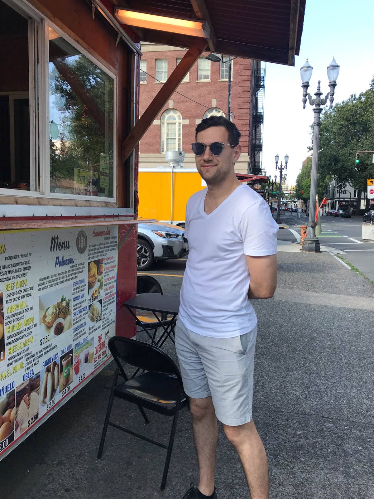 A man with short dark hair and round sunglasses in a white shirt and light shorts, standing in front of a food truck.