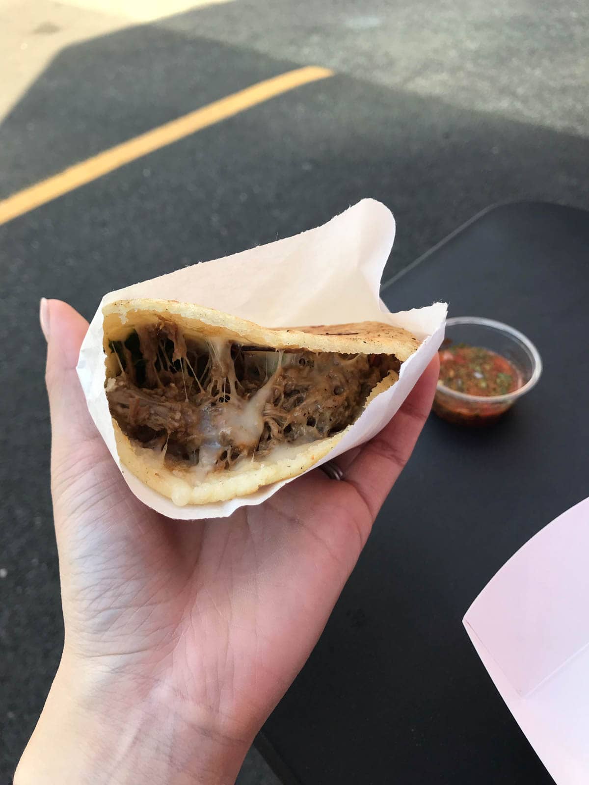 A woman’s hand holding an arepa – a tortilla with beef and cheese inside, in a white paper bag.