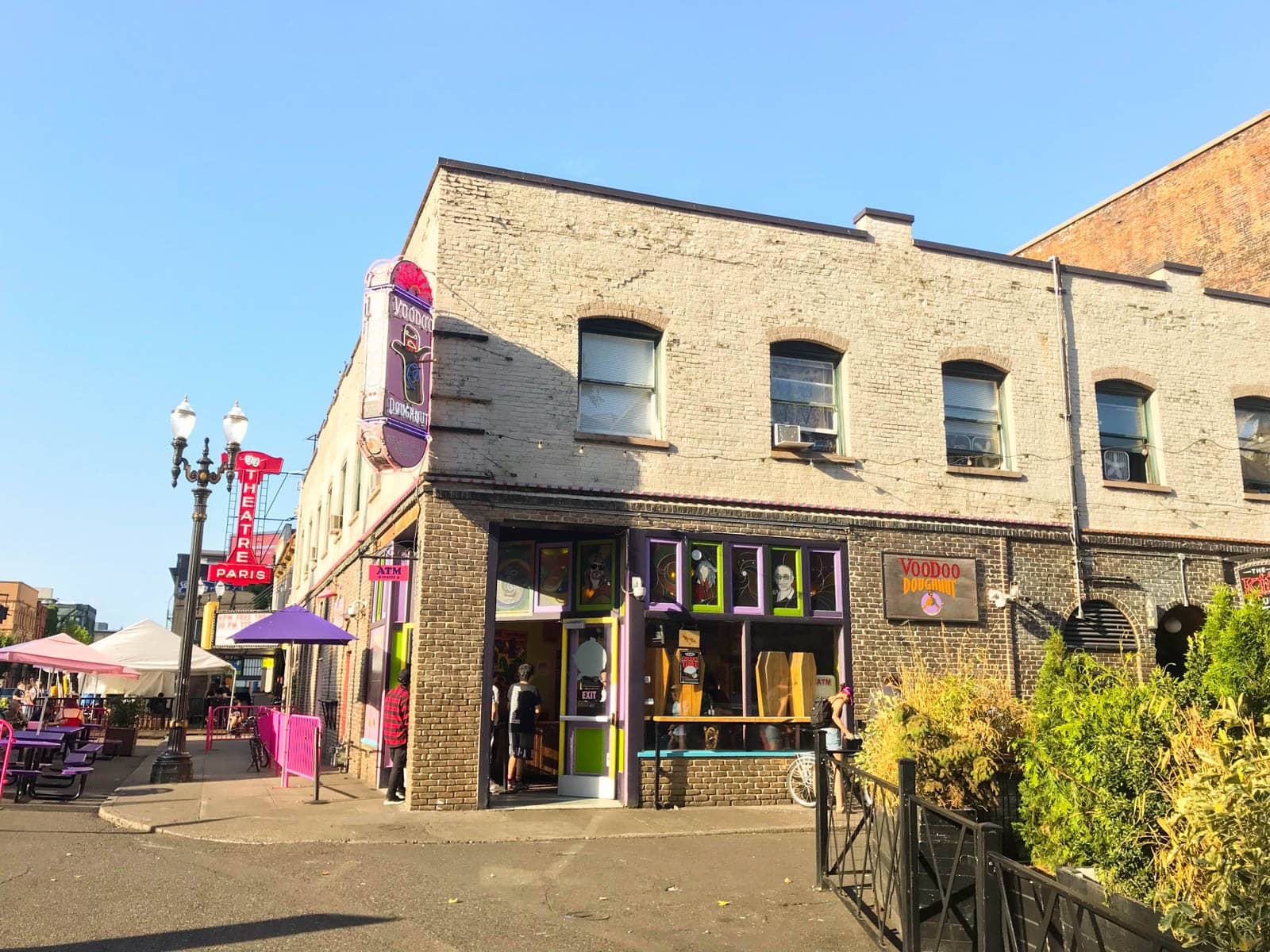 The front of a doughnut shop, decorated with brightly coloured signage in purple, pink, and lime green. A sign reads “Voodoo Doughnut”. It’s a sunny day and the sky is very blue