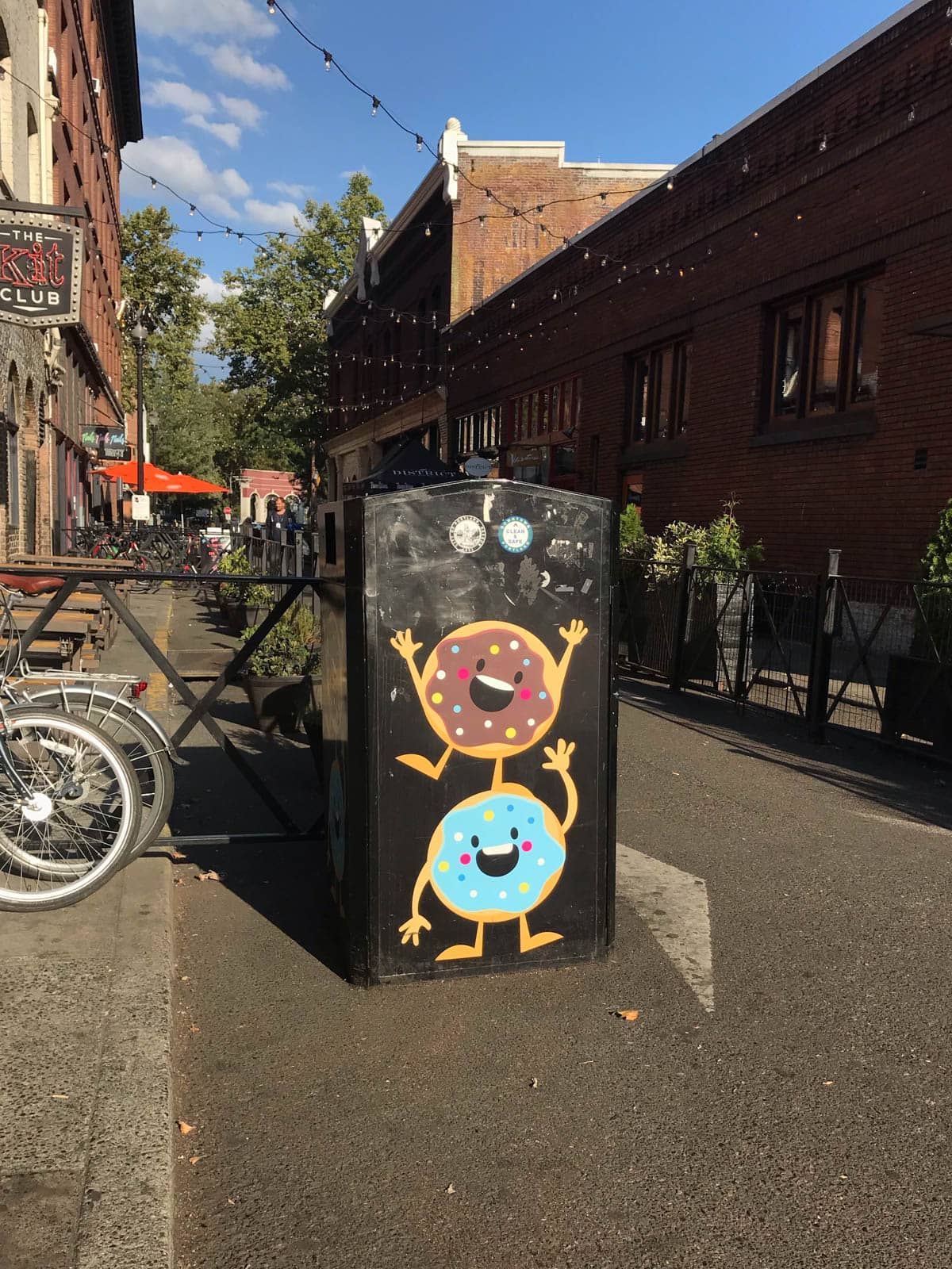 A black communal trash can in the middle of a city alleyway, with two smiling doughnuts painted on the trash can