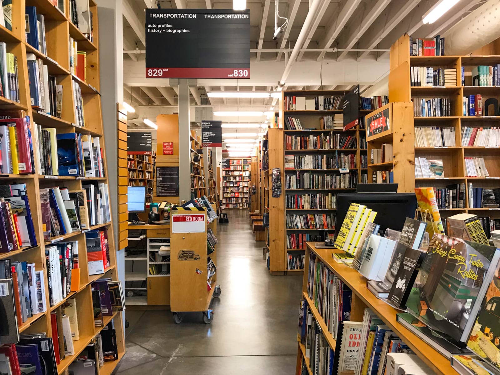 The inside of a bookstore with yellow-brown shelving, almost as tall as the ceiling, filled with books. Signs from the ceiling indicate the various genres of books in each of the aisles.