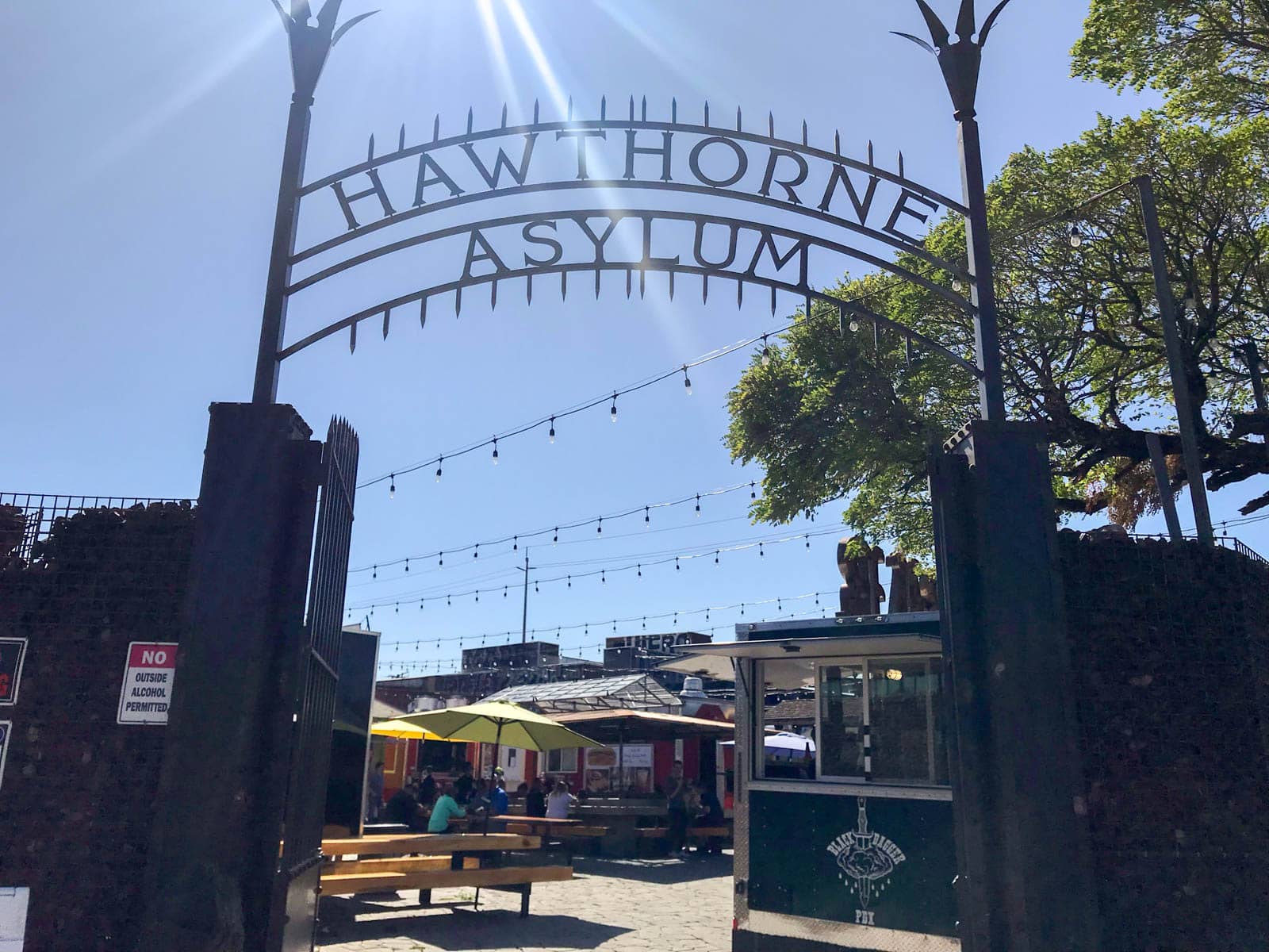 The entrance to a block of food trucks, with the gates open and the signage above the gate reading “Hawthorne Asylum”.