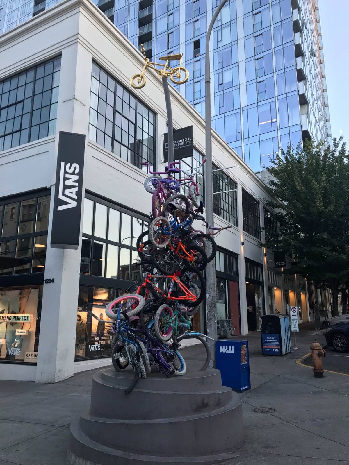 A tall structure of many small colourful bicycles atop each other, standing as a tower with a small golden bicycle on top. The structure is in the middle of a city street with some shopfronts.