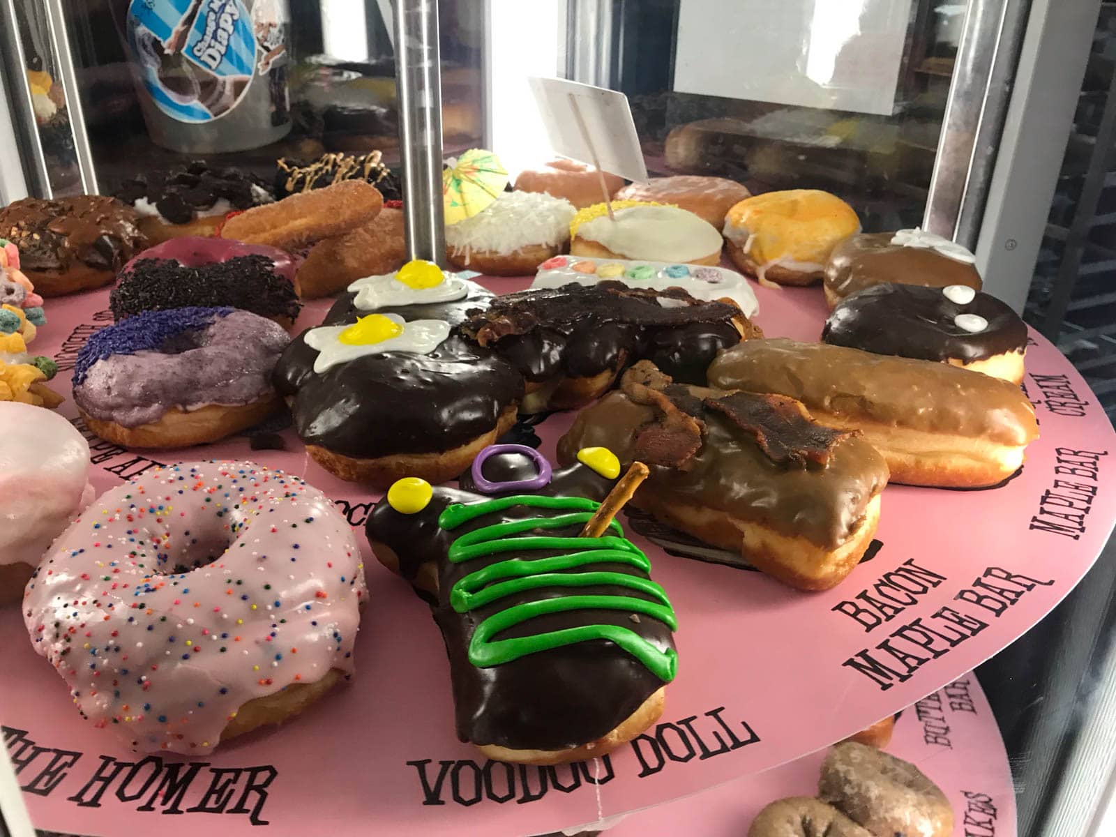 A multi-tier display with pink cardboard plates, with colourfully decorated doughnuts on them. The doughnuts are labelled by text printed on the pink cardboard.