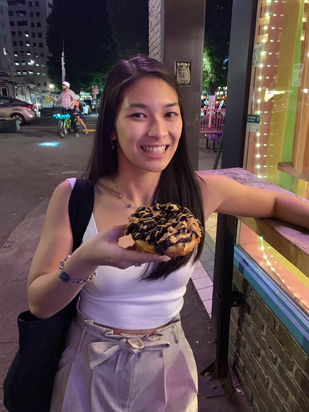 A woman with long dark hair, holding a doughnut topped with chocolate biscuits and peanut butter sauce