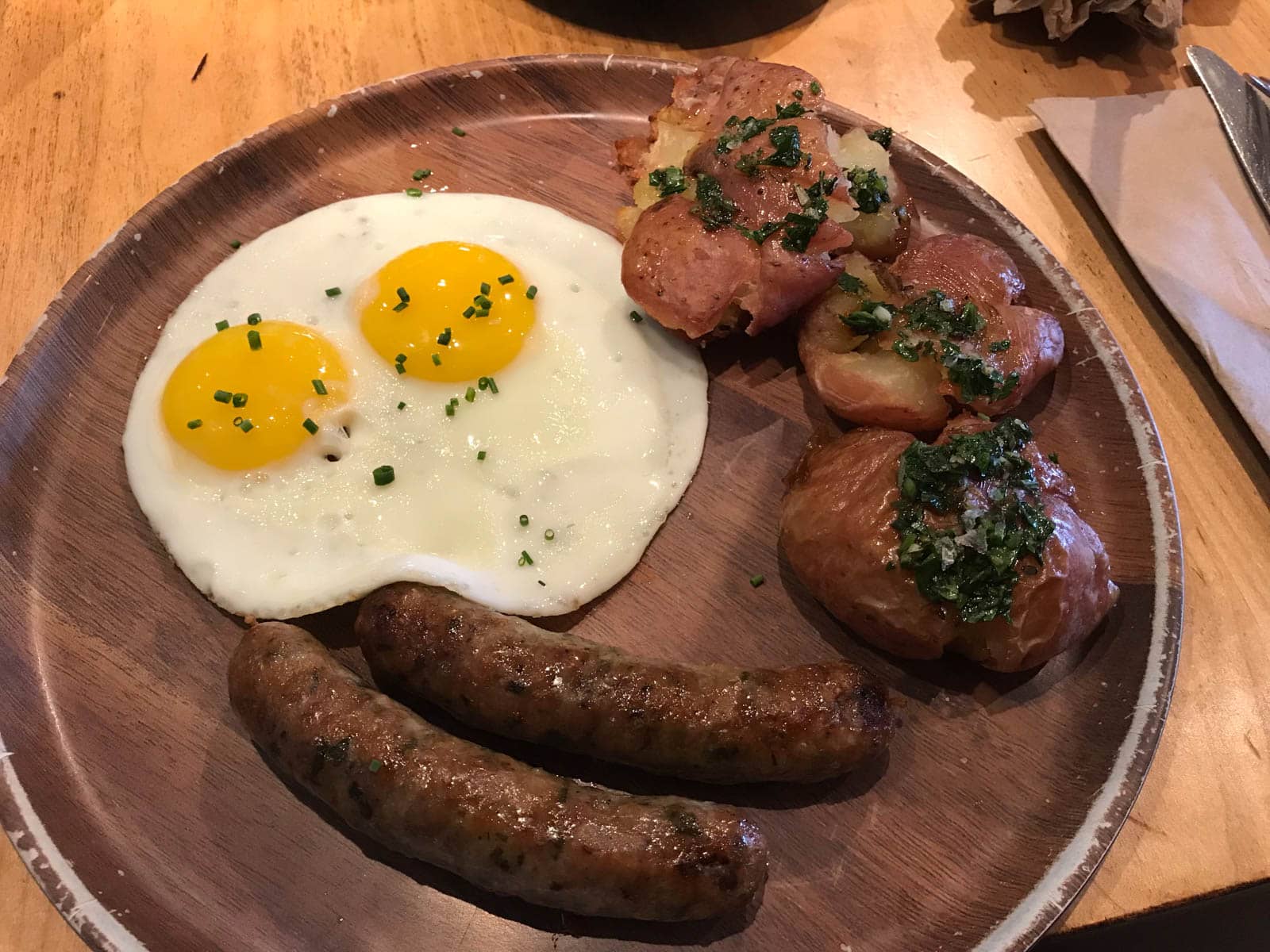 A round wooden plate served with perfect fried eggs, baked potato and two sausages