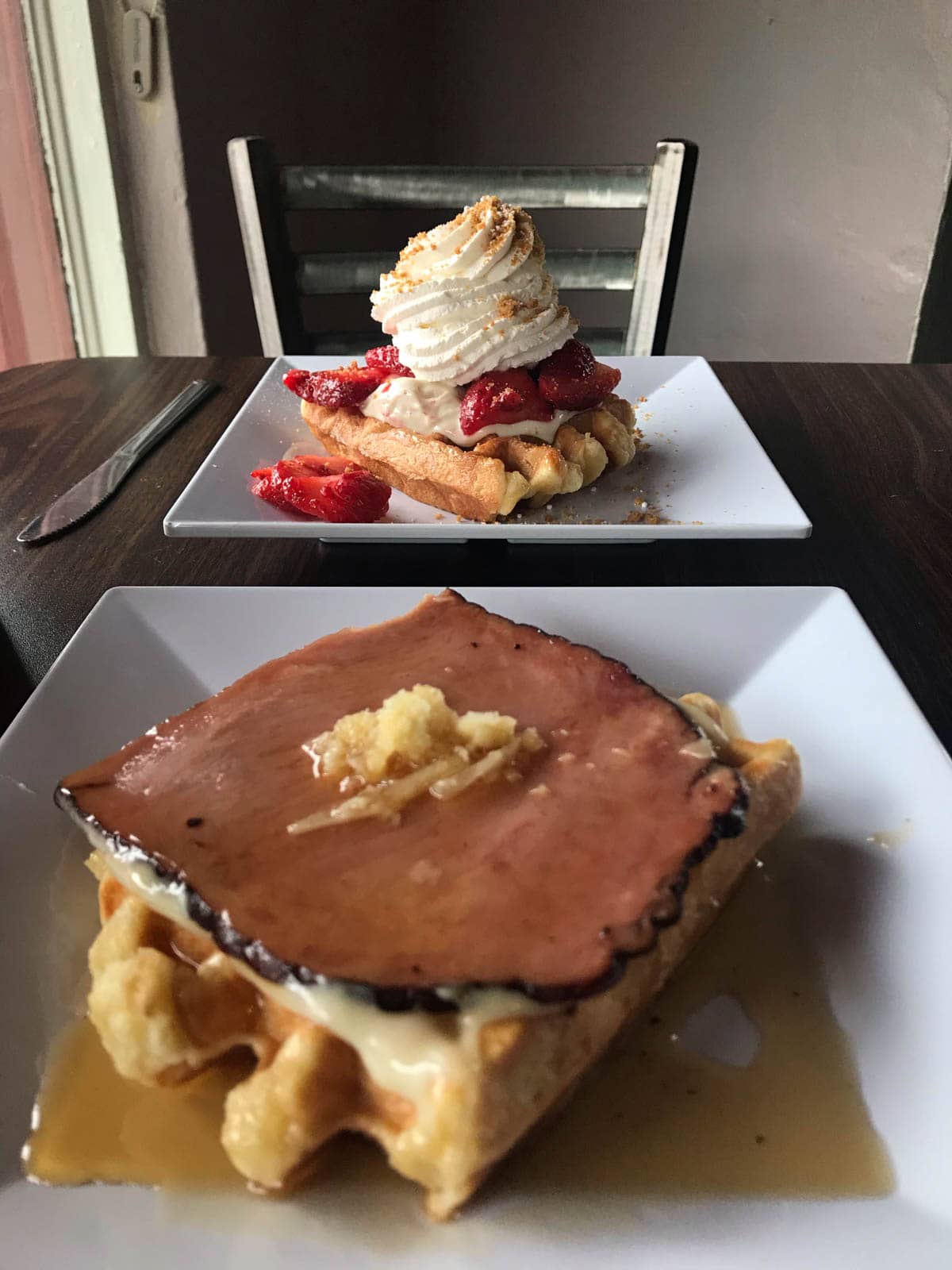 Two waffles served on square dishes. One is topped with ham and cheese, and the one in the background is topped with strawberries and whipped cream.