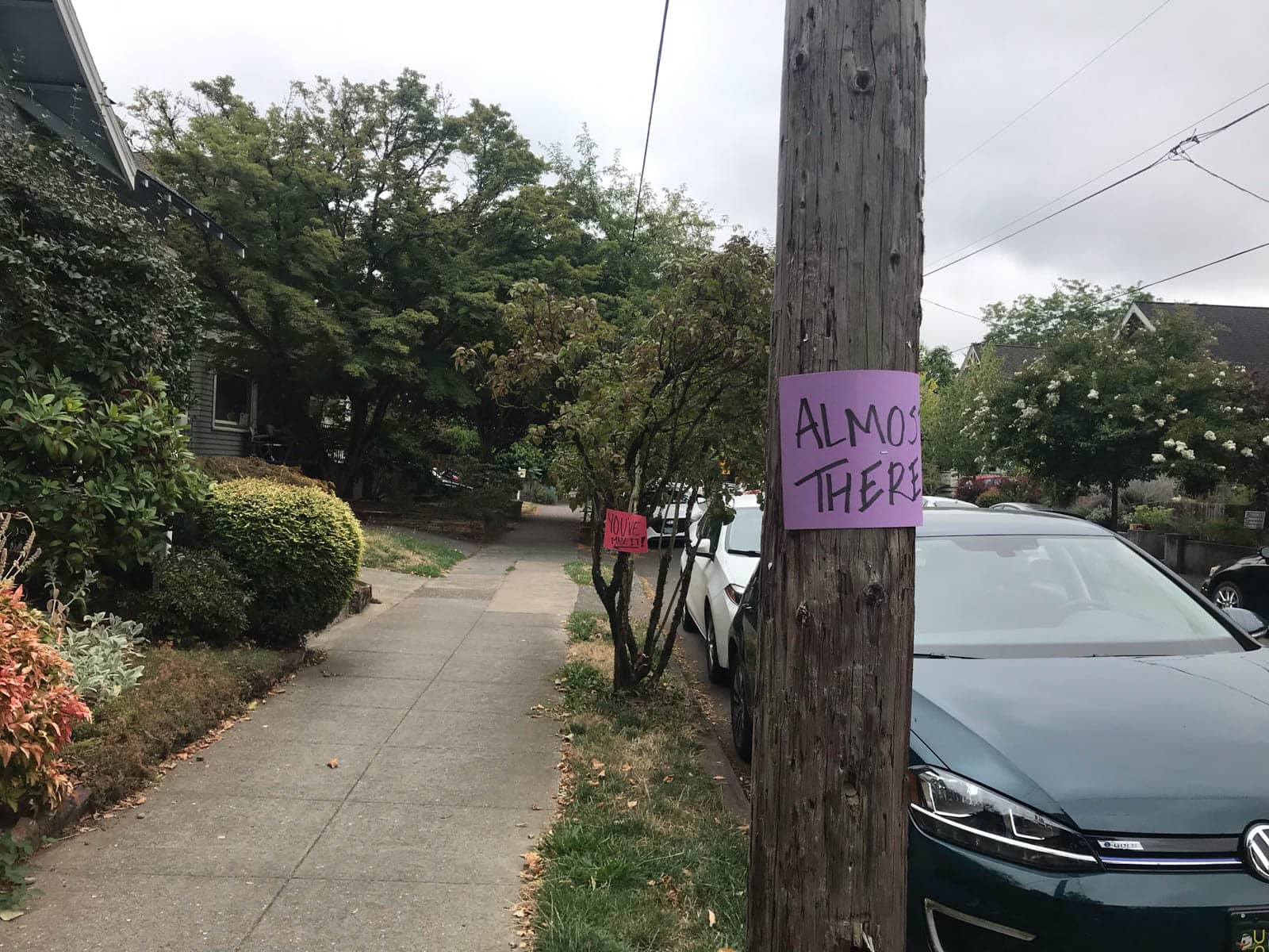 A path on the side of a suburban street. The street pole has a purple piece of paper with “almost there” written on it”, and further away, a tree with a red sign reading “you’ve made it!”