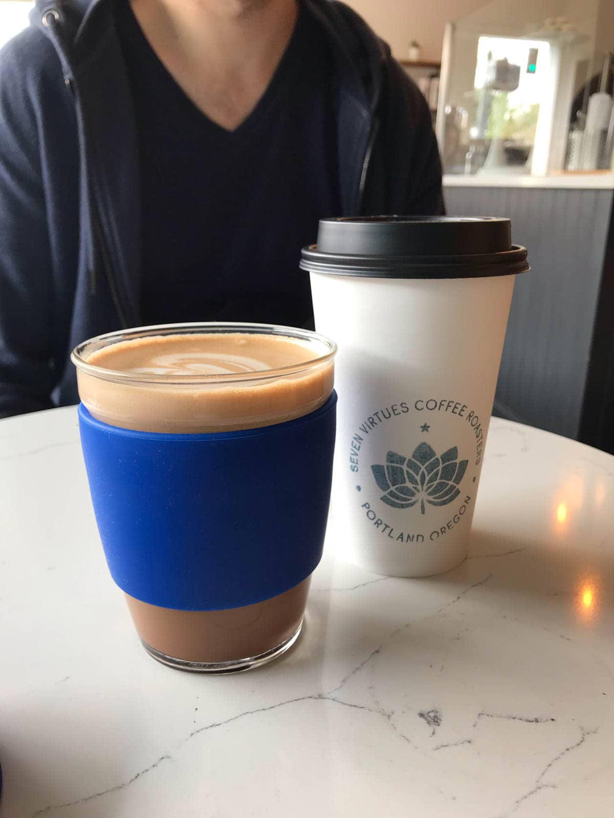 A glass coffee cup with a blue rubber grip, filled with coffee, alongside a paper cup reading “Seven Virtues Coffee”.