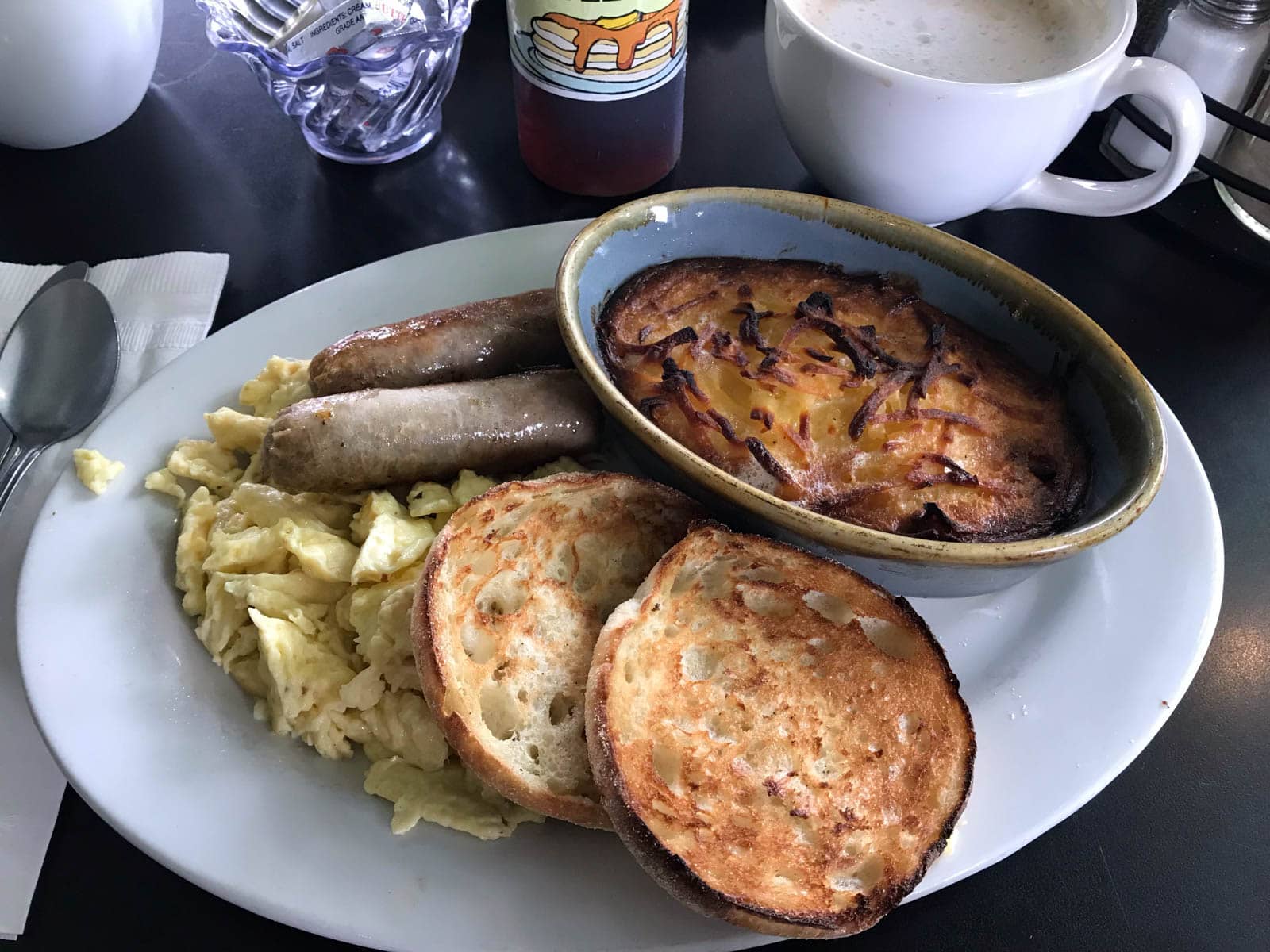 A white oval dish with sausages, scrambled eggs, a toasted muffin and potato rosti served on it.