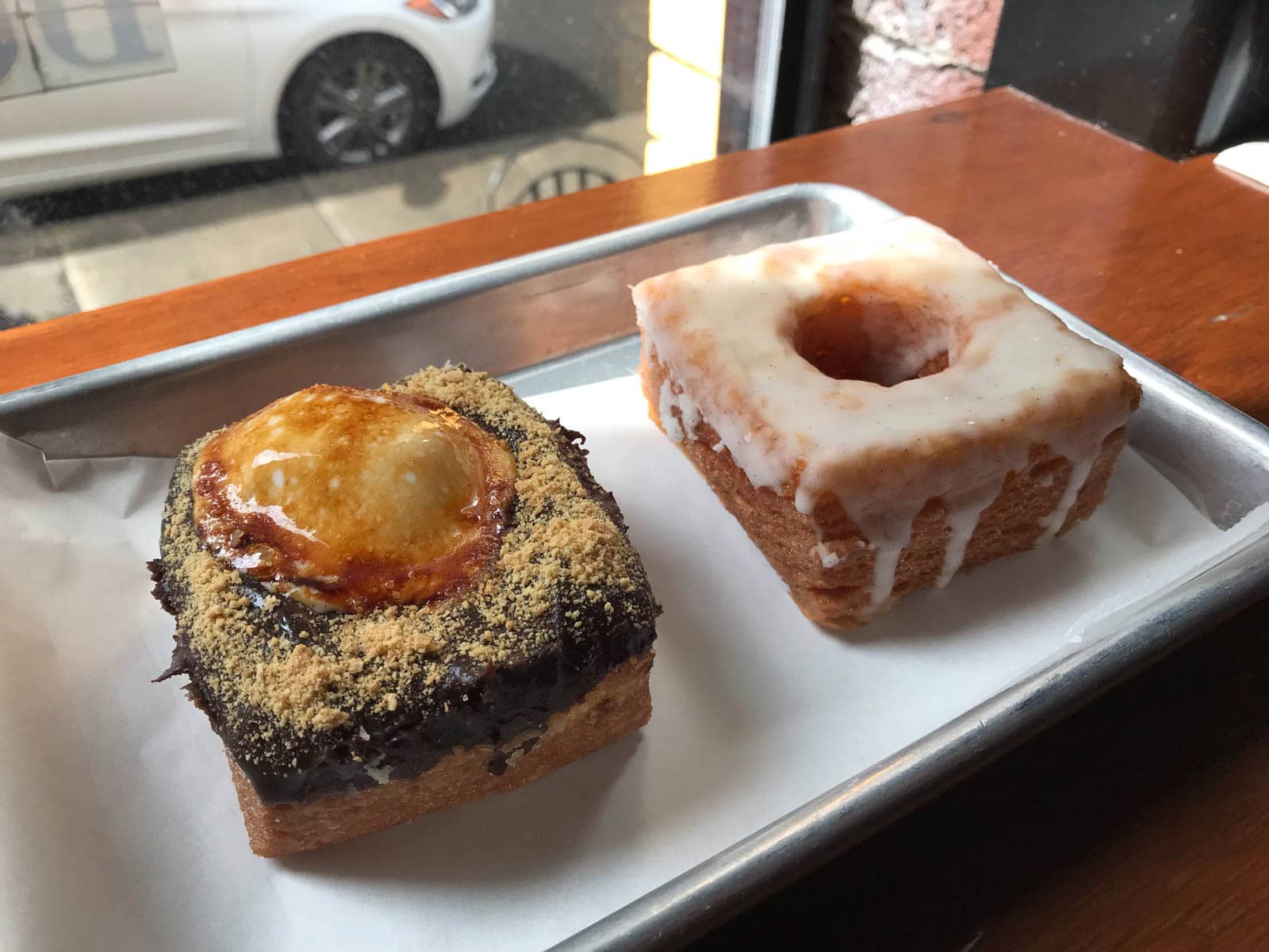 Two square cronuts (croissant doughnuts) in a silver tray. One has a white frosted glaze while the other one has a chocolate and caramel topping