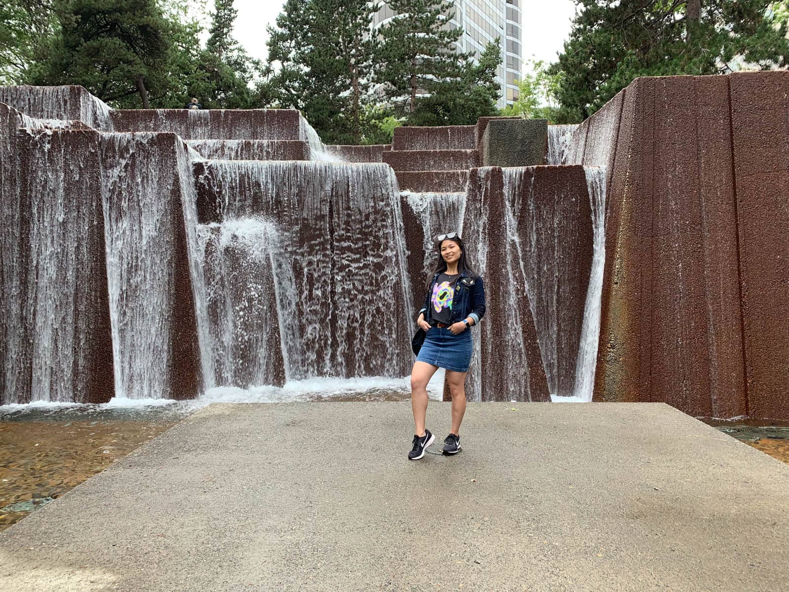 A woman in a denim jacket and denim skirt with her hands in her pockets, standing in front of large stone rocks with waterfalls running down them.