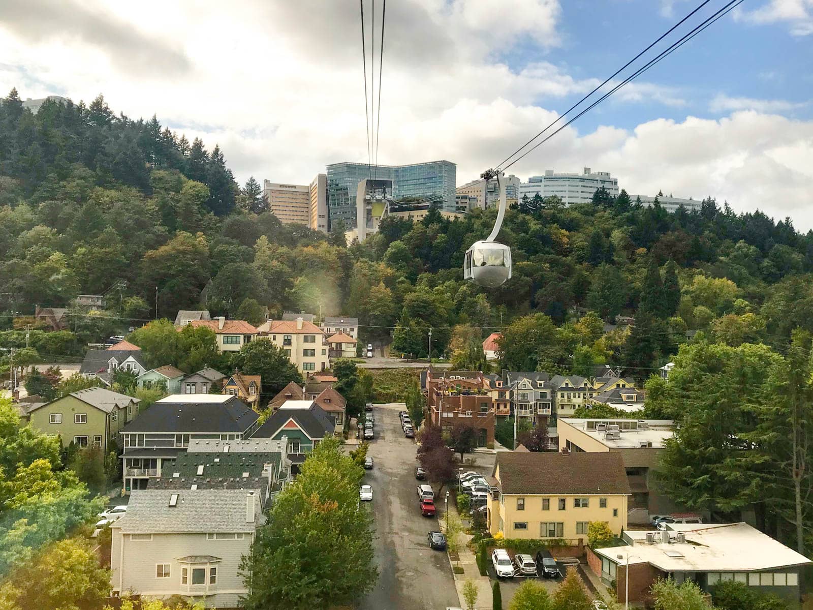 A flew from an aerial tram, where one of the other carriages can be seen. The aerial tram cables are going uphill. Residential houses sit below the tram, and the sky is blue but filled with many clouds. Towards the top of the hill is a lot of vegetation.