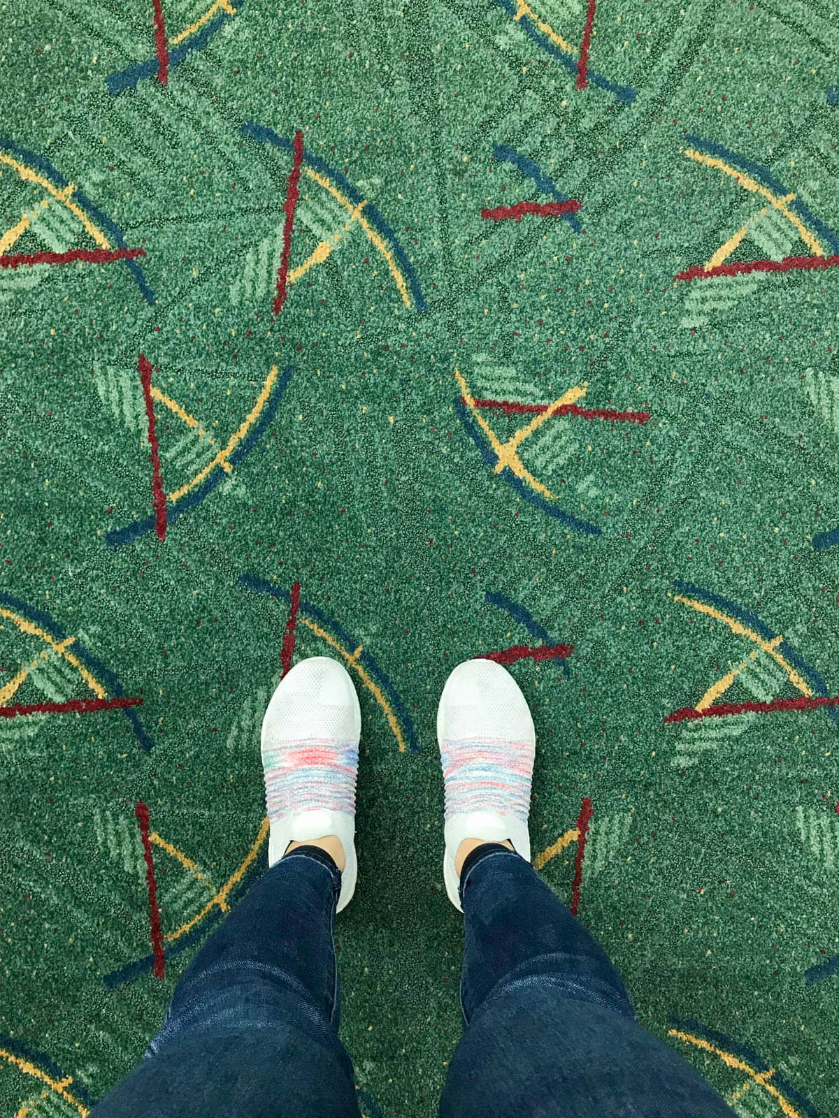 A top-down view of green patterned carpet; the white sneakers of the woman taking the photo can be seen.
