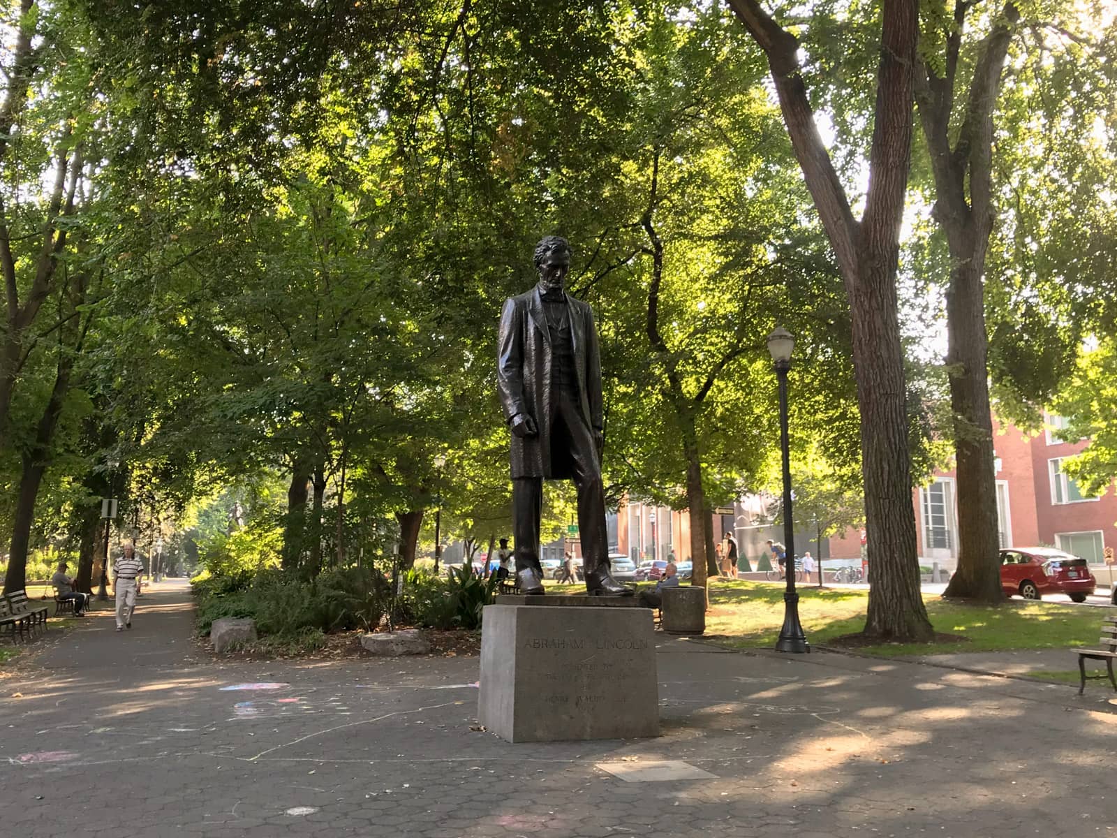 A statue of a man on top of a block with an engraving, in a park with many trees