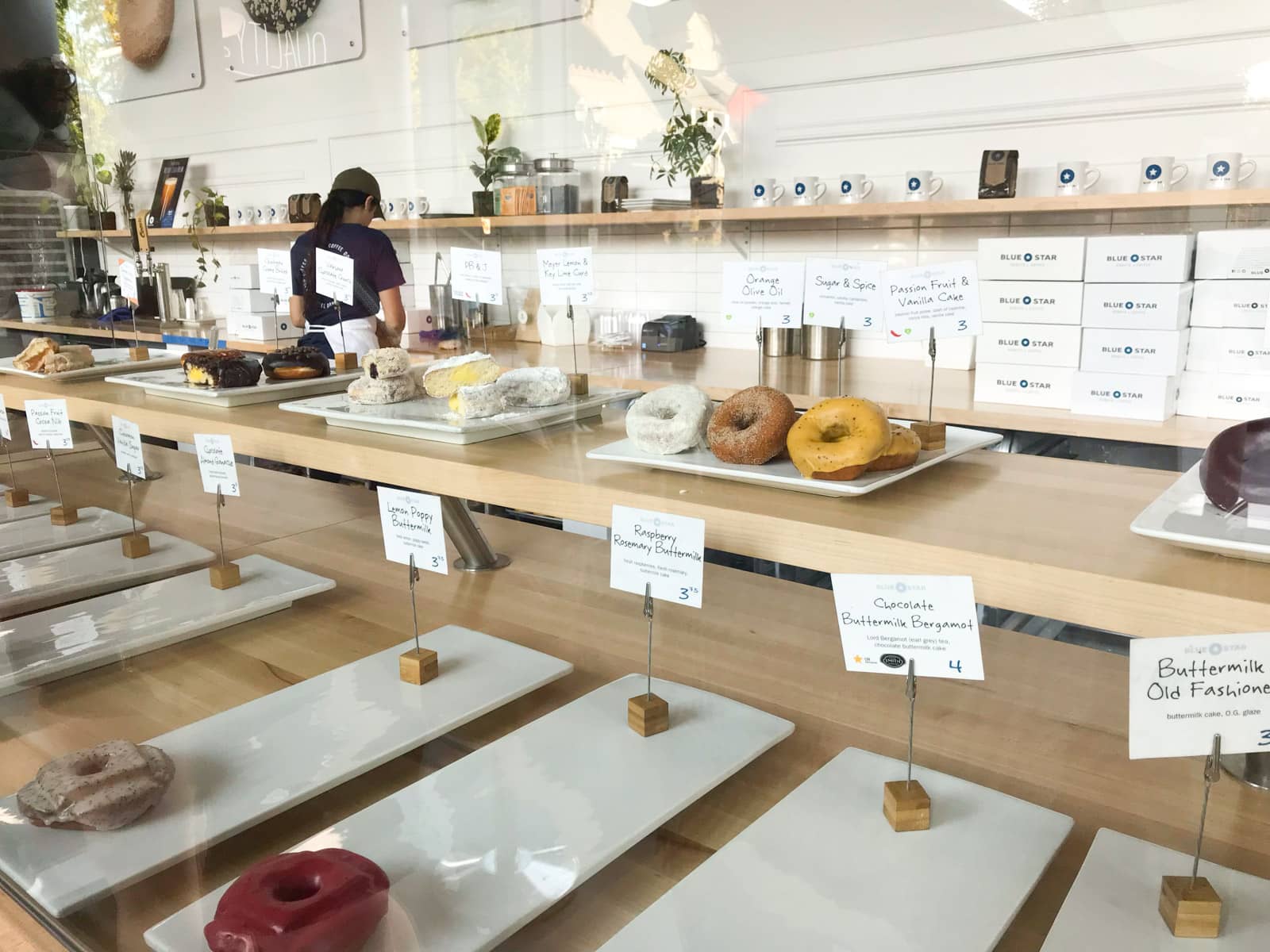 The inside of a doughnut shop, with many gourmet doughnuts on display on long white trays.