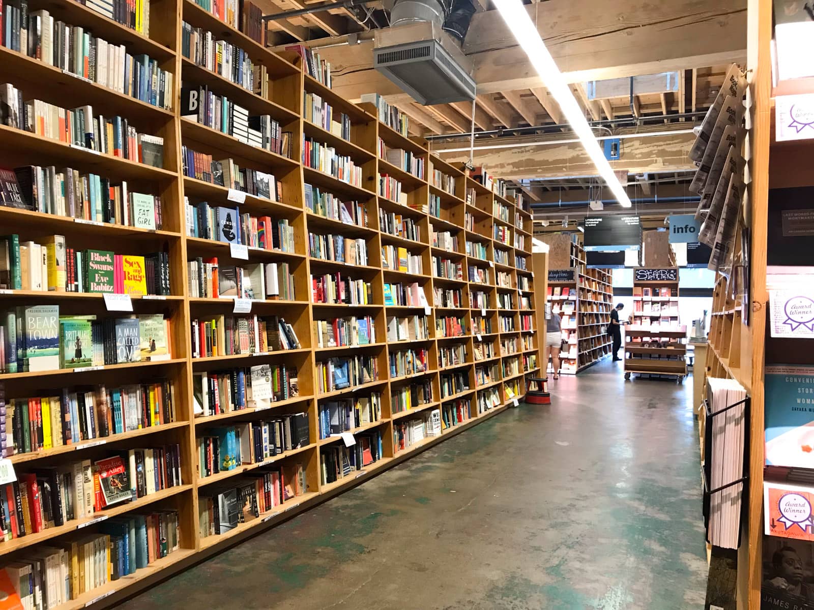 The aisle of a large bookstore, with wooden shelves as tall the ceiling. All the shelves are full of books