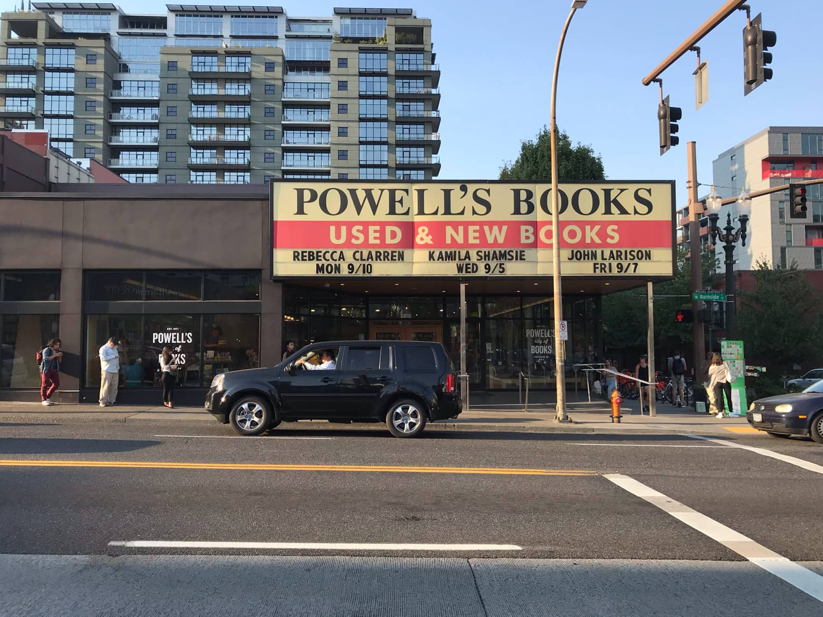 The front of a bookstore seen from the opposite side of a street. The signage reads “Powell’s Books – Used & new books”. A couple of cars can be seen driving past. There are a few people waiting in front of the bookstore.