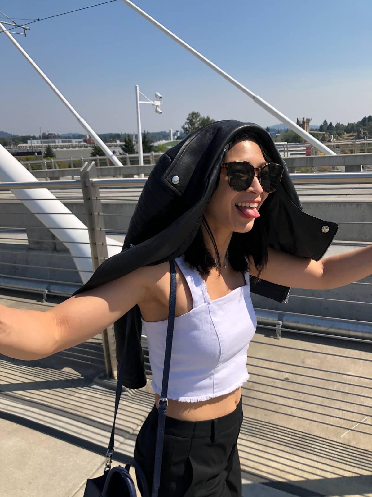 A woman in a white denim crop top and black pants, with big and round dark sunglasses and a leather jacket on top of her head like a cape. She is walking on what appears to be a bridge and is making a face with her tongue slightly stuck out.