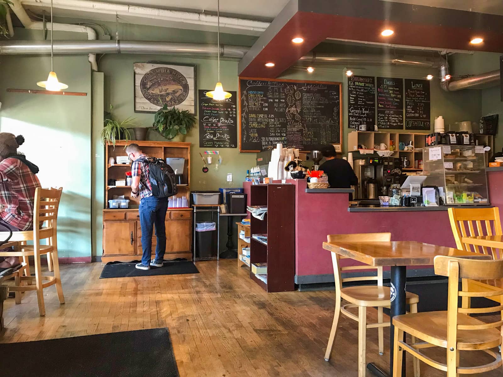 The inside of a coffee shop. The counter can be seen, with chalkboards with handwritten menus. There are only a few people in the cafe and a few empty chairs. The decor is brown and muted and gives a homey vibe.