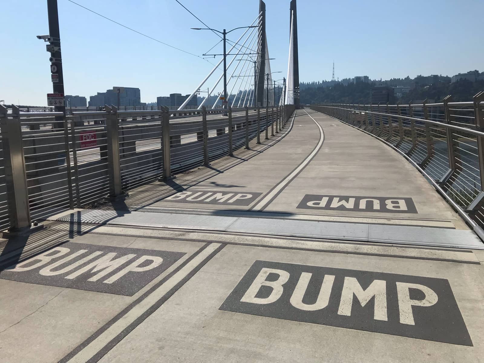 The wide walkable path on a bridge with metal railings. Markings on the floor of the bridge read “Bump” to indicate a small bump in the path for pedestrians