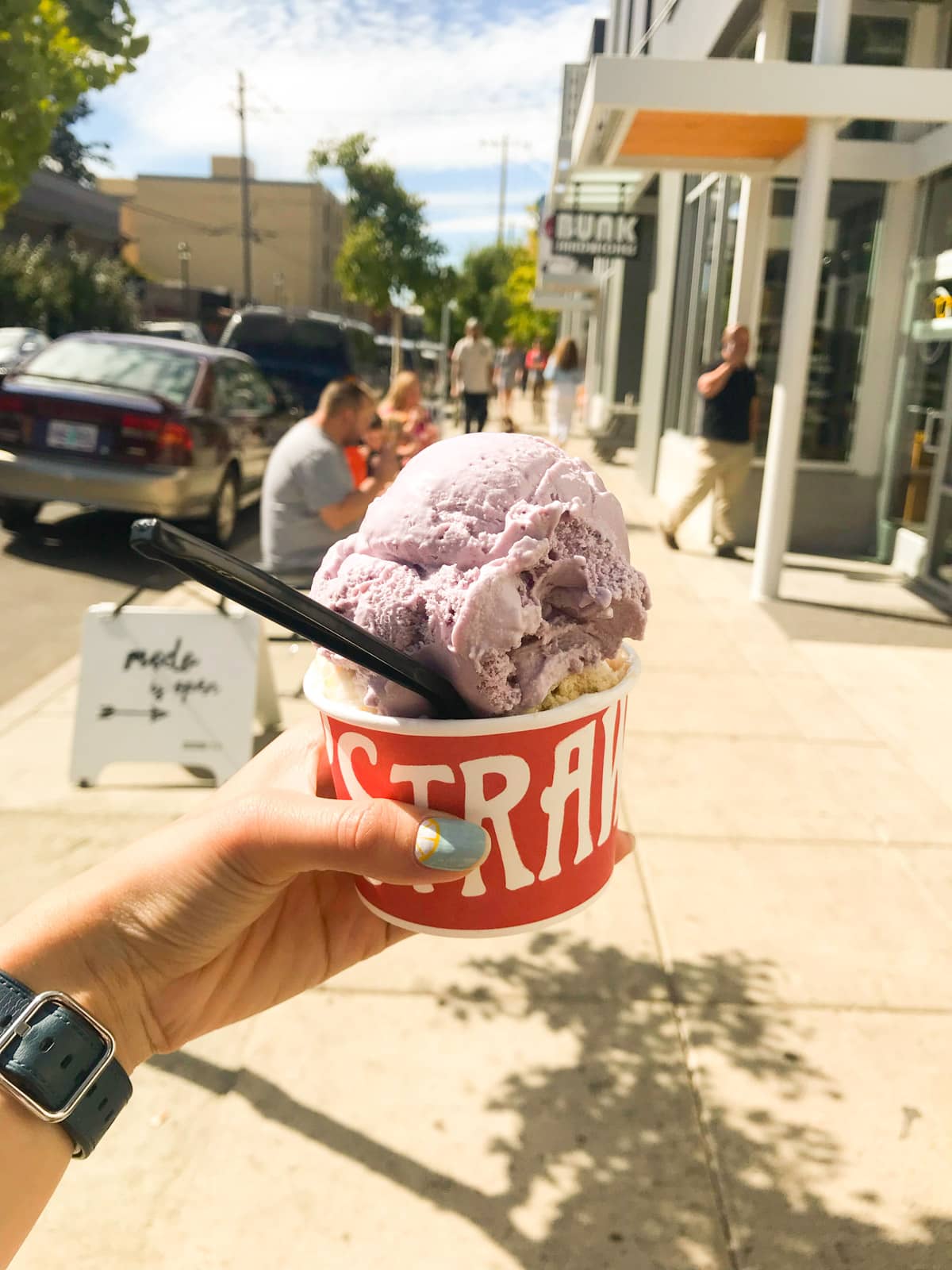 A woman’s hand holding a red-and-white paper cup with a large serving of ice cream. The top scoop is light purple and the one underneath can only just be seen, and is beige in colour.