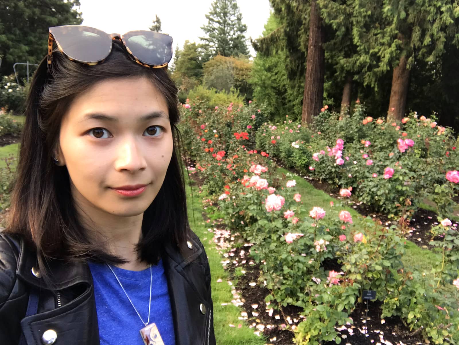 A selfie of a woman with short dark hair, wearing a blue shirt and black jacket. She is in the middle of a rose garden