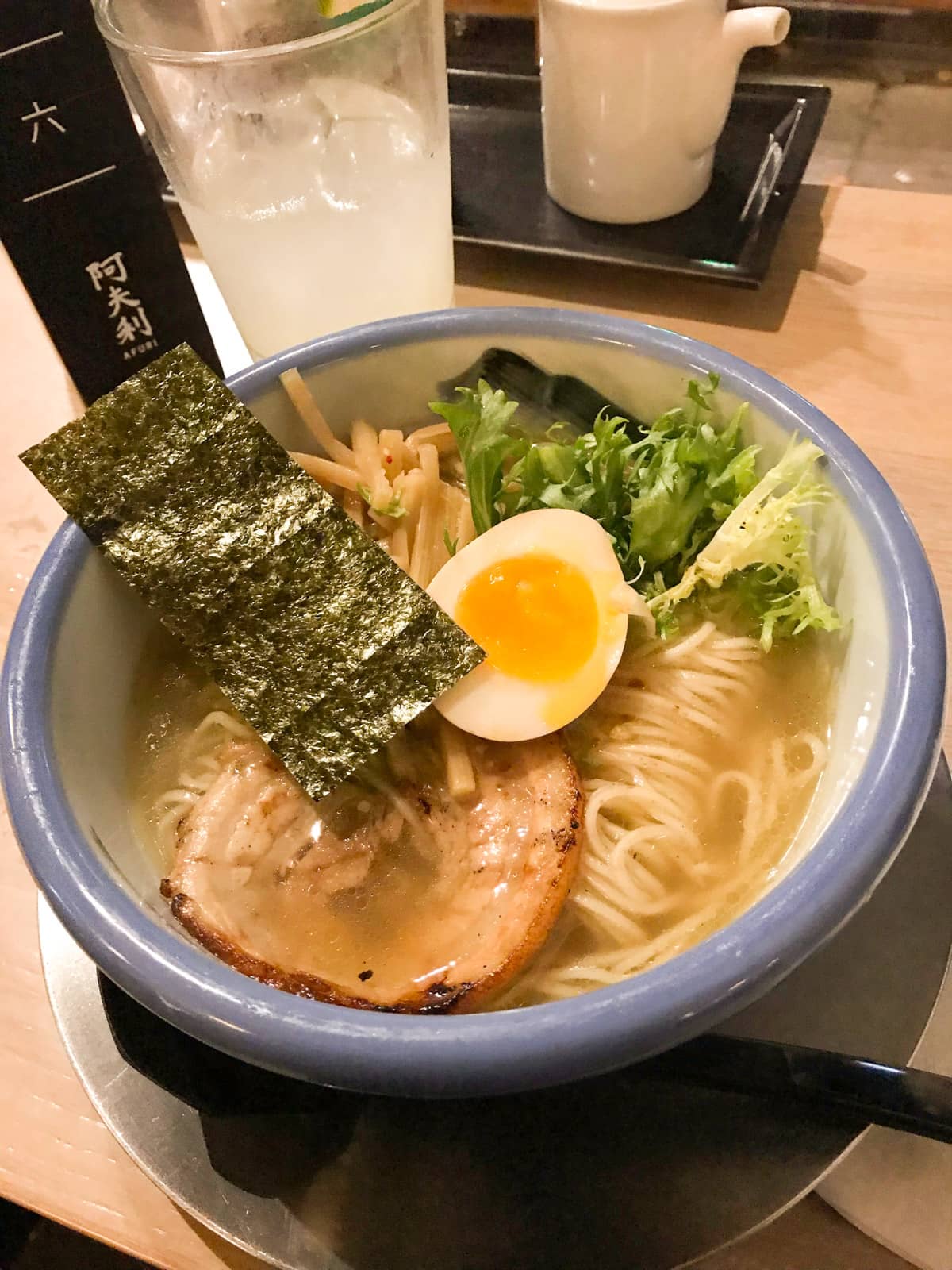 A blue-rimmed bowl filled with pork ramen. It is topped with half a boiled egg, bamboo shoots and nori seaweed