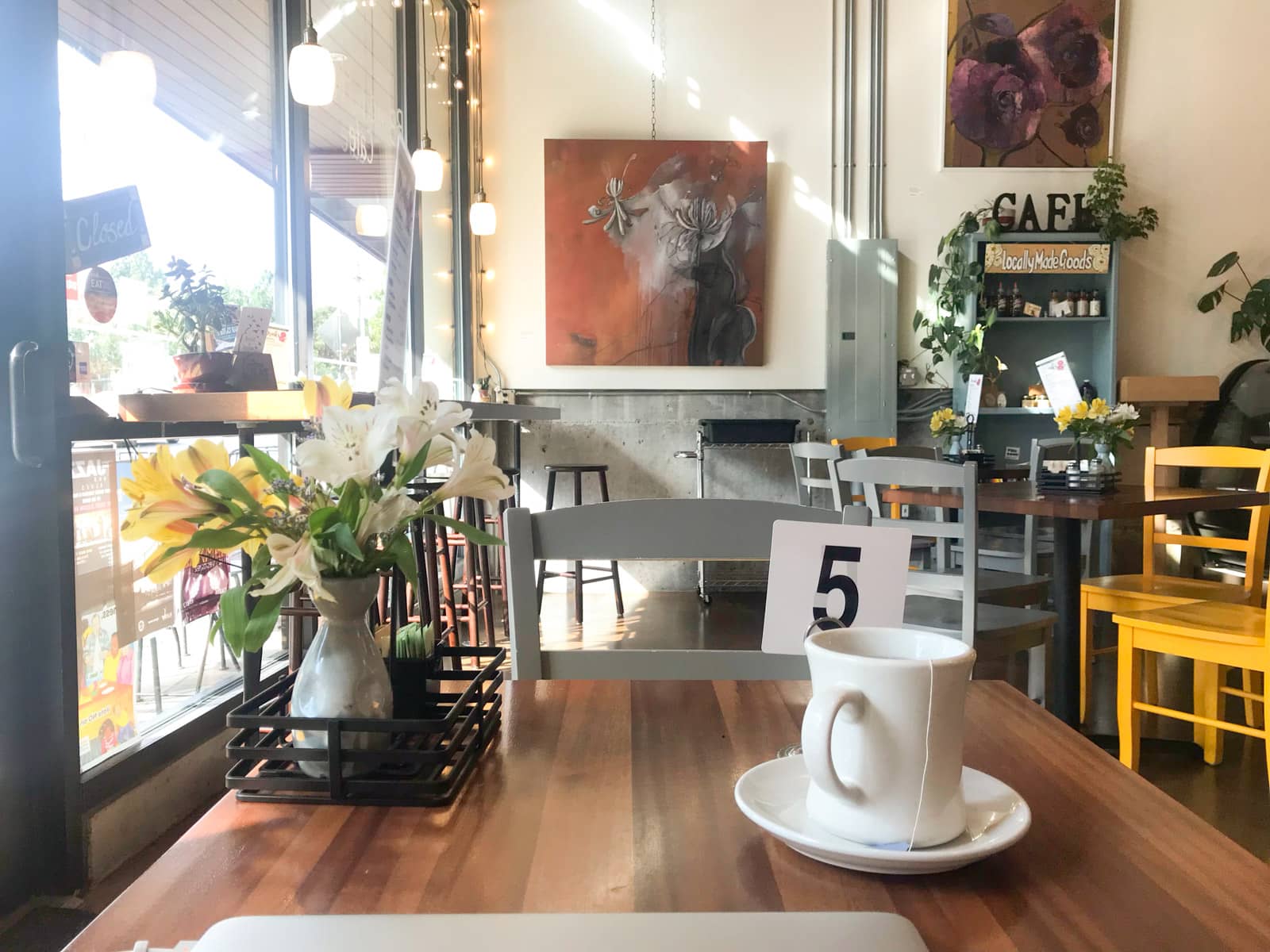 The inside of a cafe seen from one of the tables inside. The table has a table number and a cup of tea. The cafe has some art on the walls and some yellow wooden chairs.