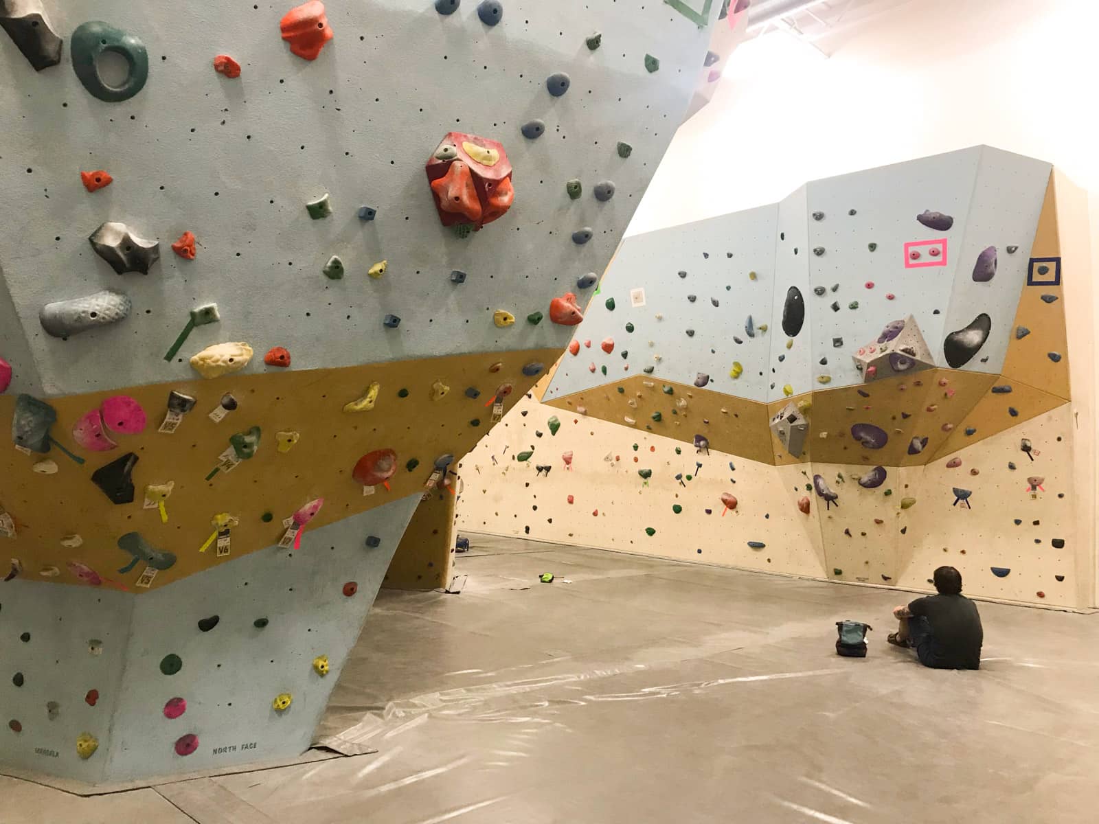 The inside of a bouldering/rock climbing gym. Coloured rock holds line the geometrically-shaped walls, and someone sits on the landing, facing away from the camera.