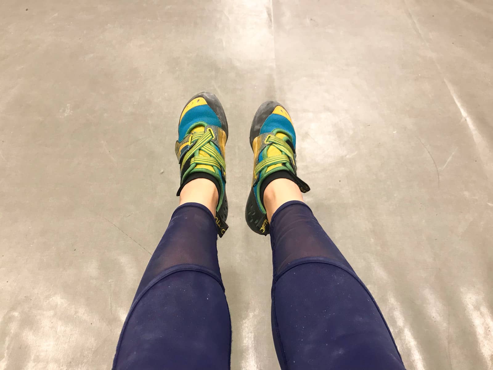 A top-down view of a woman’s feet; she is wearing bright yellow and blue rock climbing shoes, and navy blue leggings.