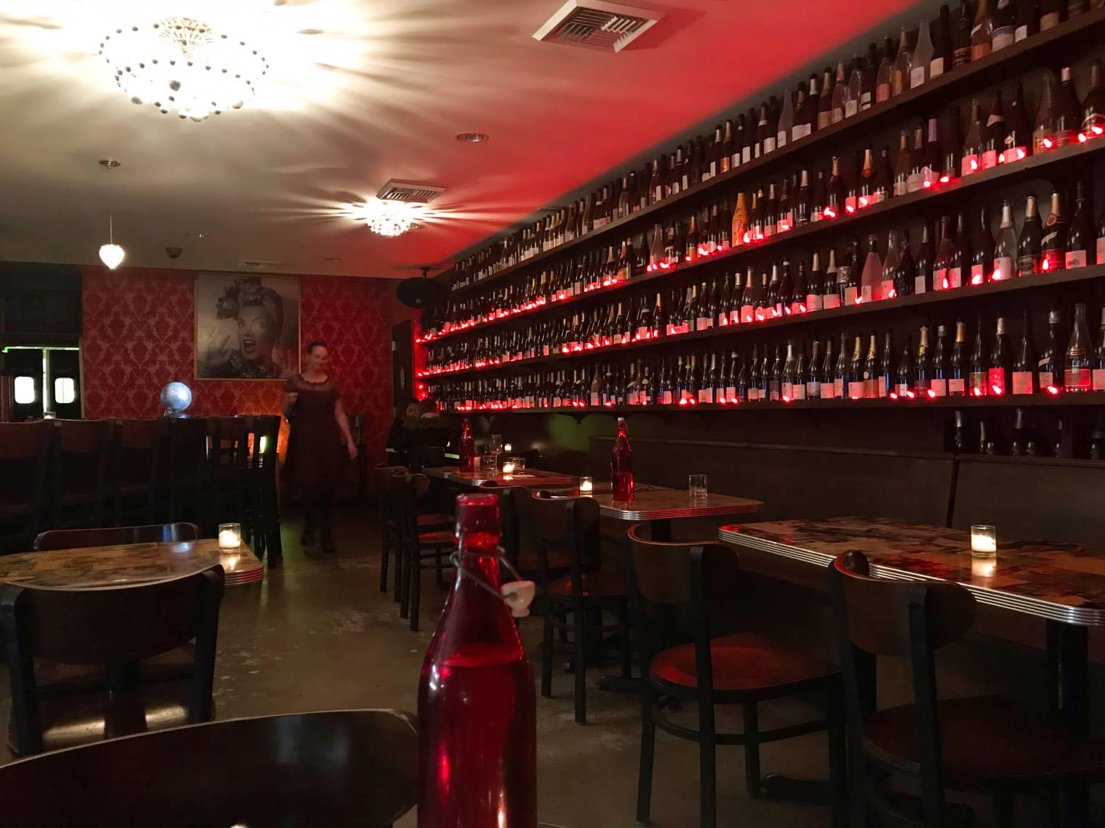 The dark and dimly lit interior of a cafe, with red decor. Wine bottles line the walls along with fake red candles.