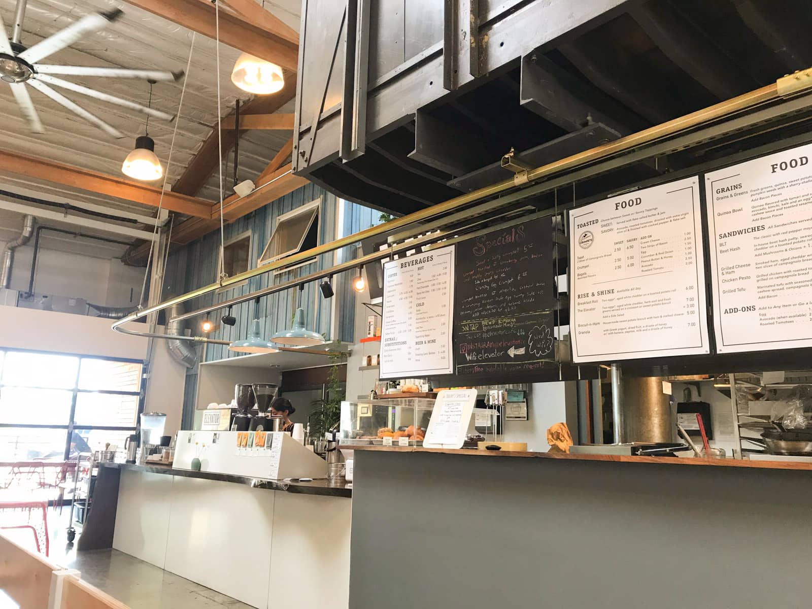 A cafe counter with a menu above the counter. Hanging lighting and fans can be seen from the ceiling.