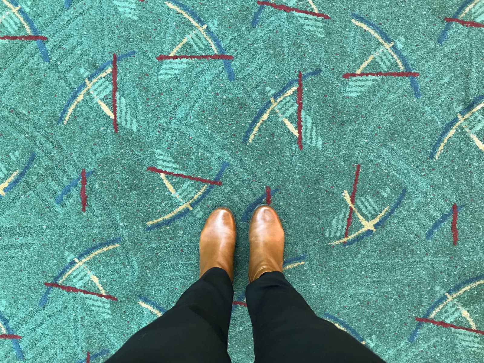 A top-down view of a woman with brown boots on her feet. She is standing on a teal green carpet with a repeating pattern