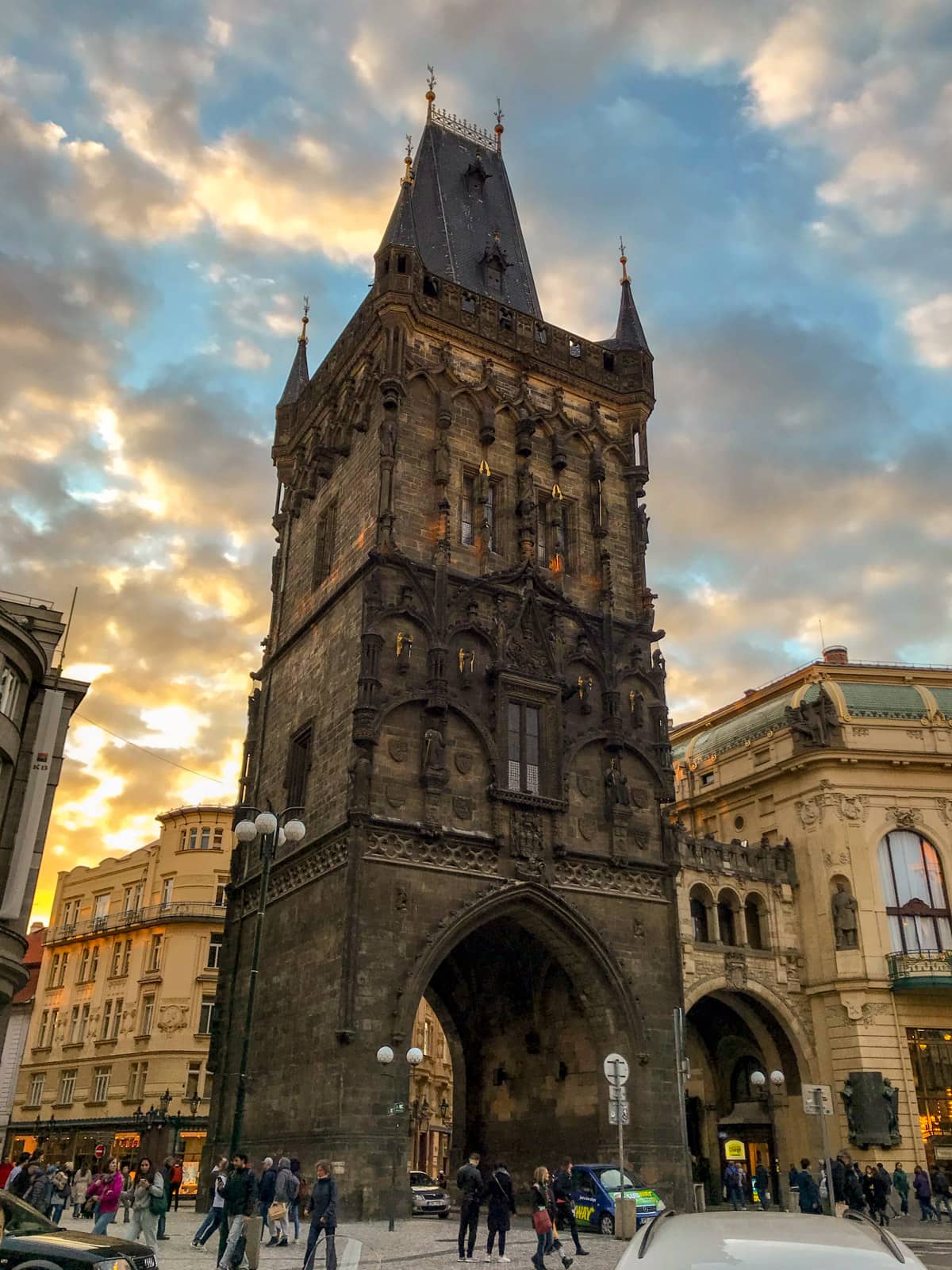 A tower with an arch underneath it to allow vehicles to pass through the road underneath. The tower is made of dark brown bricks. The sky in the background is blue with some yellow, and there are many clouds, as it’s sunset