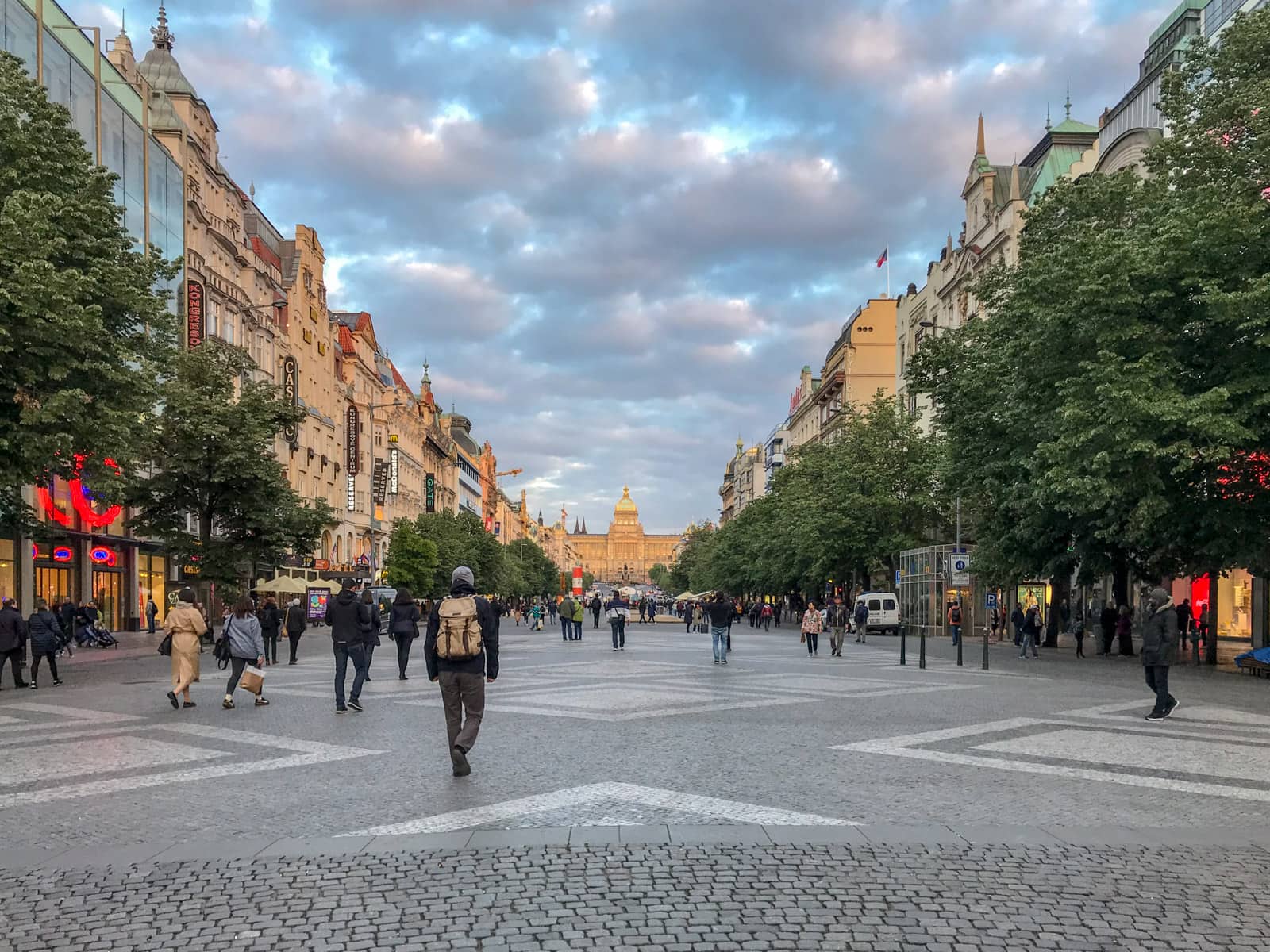 An open pedestrianised road at dusk, where the sky is a little blue-purple. There are some trees lining the sides of the road, and old buildings outside of those trees. In the distance a building can be seen centre of frame