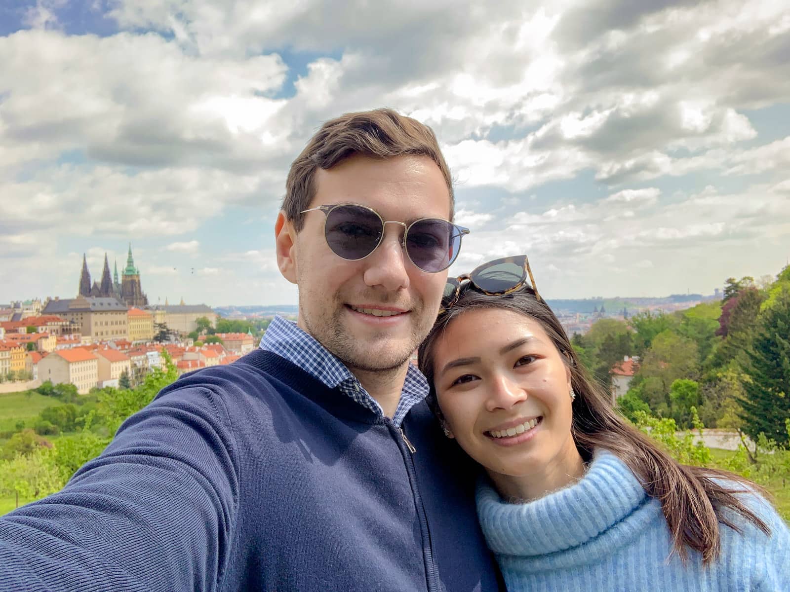 A man and woman taking a selfie, with the city of Prague in the background. The man has sunglasses on, and is wearing a dark jacket, the woman has sunglasses on top of her head and is wearing a light blue sweater.