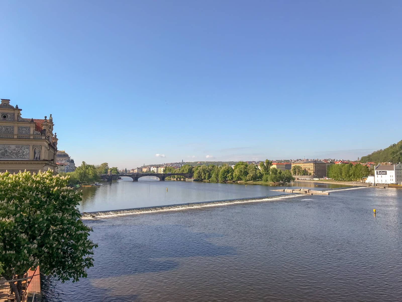 A view across a river. There is an old bridge in the distance, and a lot of green trees on the banks