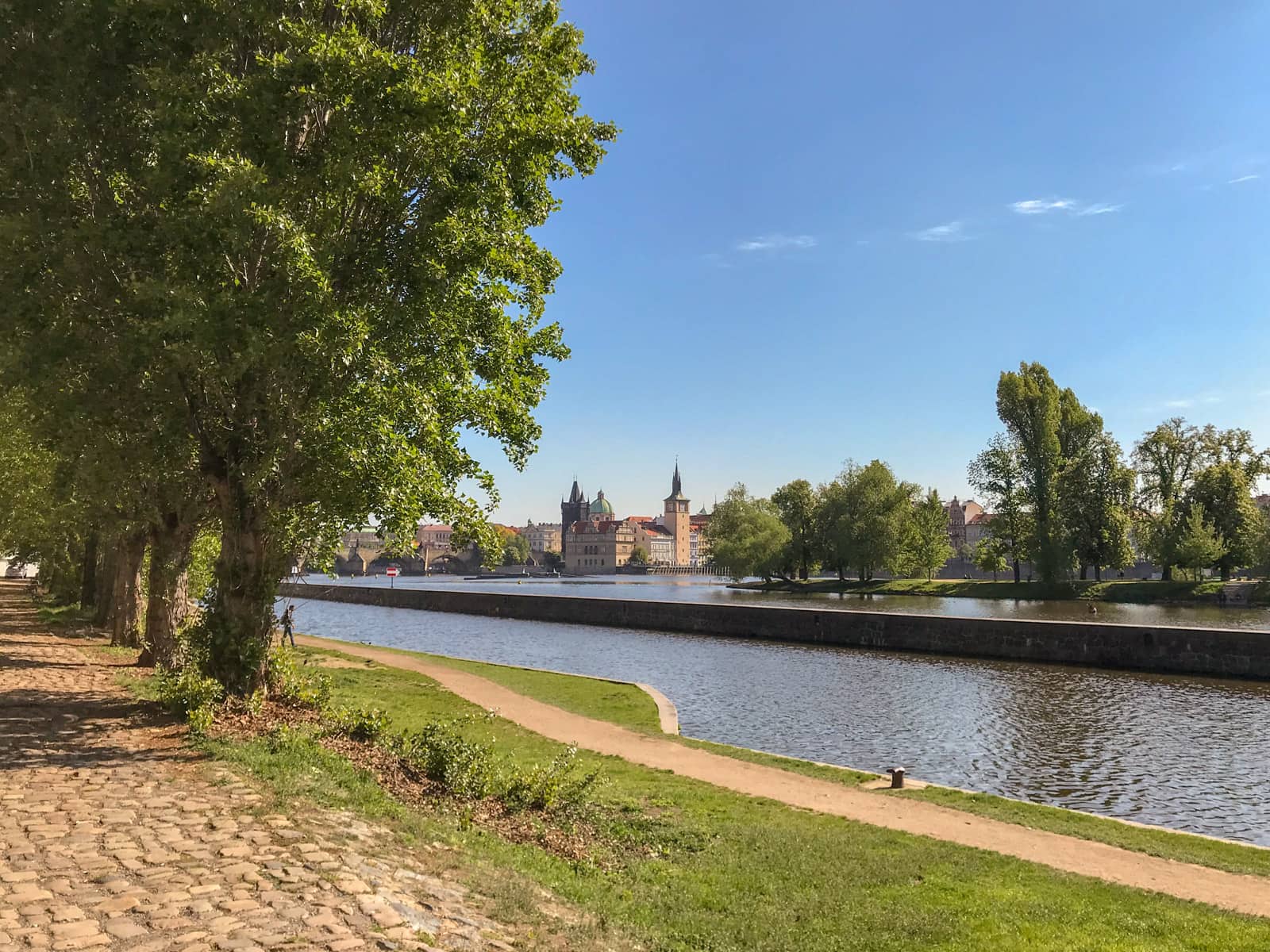 The edge of a riverbank with a path and green grass in the foreground. There are also some trees. In the distance on the other side of the river are old buildings