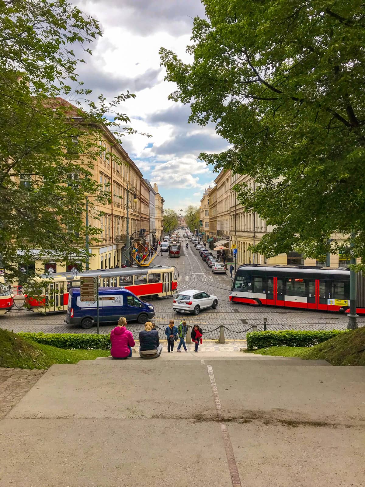 An intersection of traffic seen from a small set of stairs. There are two trams, a van and a car attempting to enter the intersection