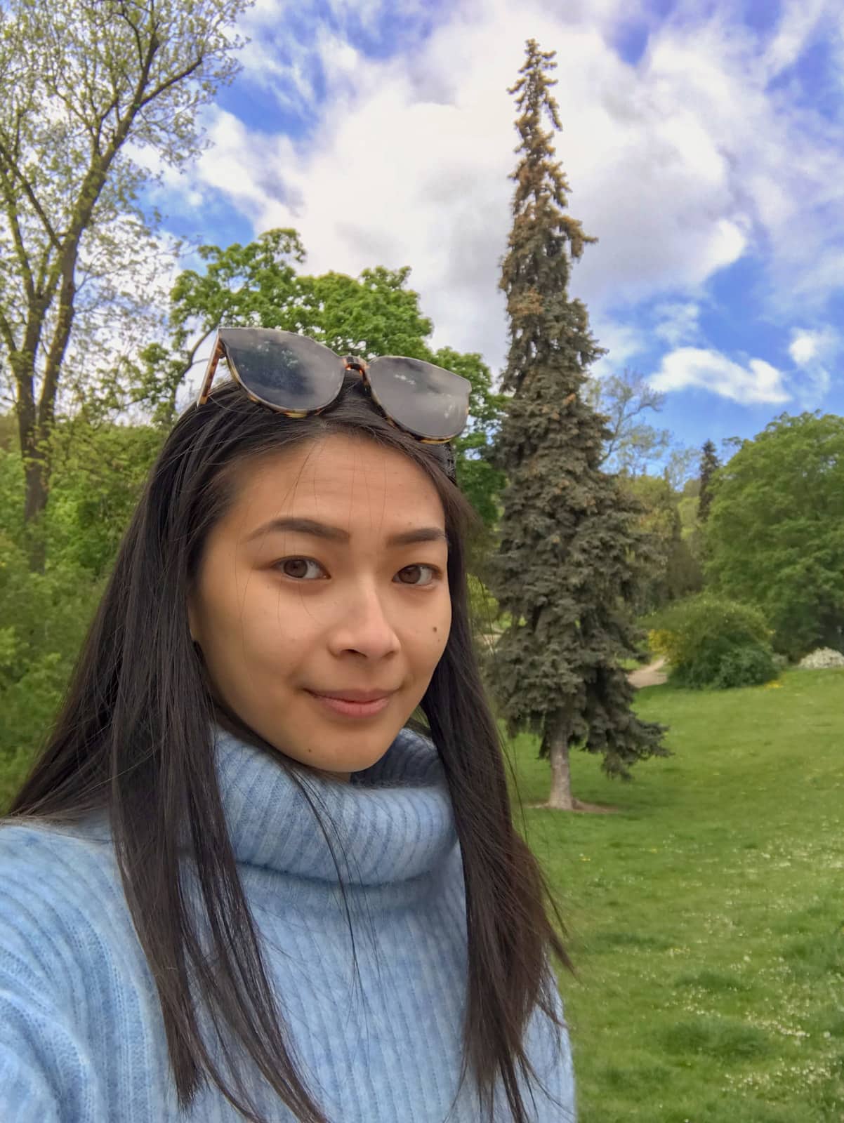 A woman with dark hair and a light blue sweater taking a selfie in an open green grassy area with lots of trees.