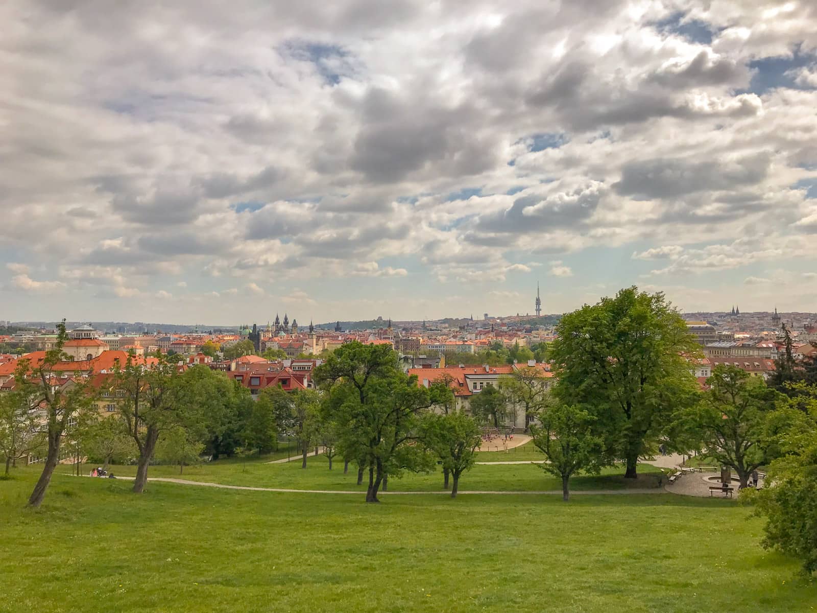 A view of Prague with a wide area of green in the foreground. There are many clouds I the sky