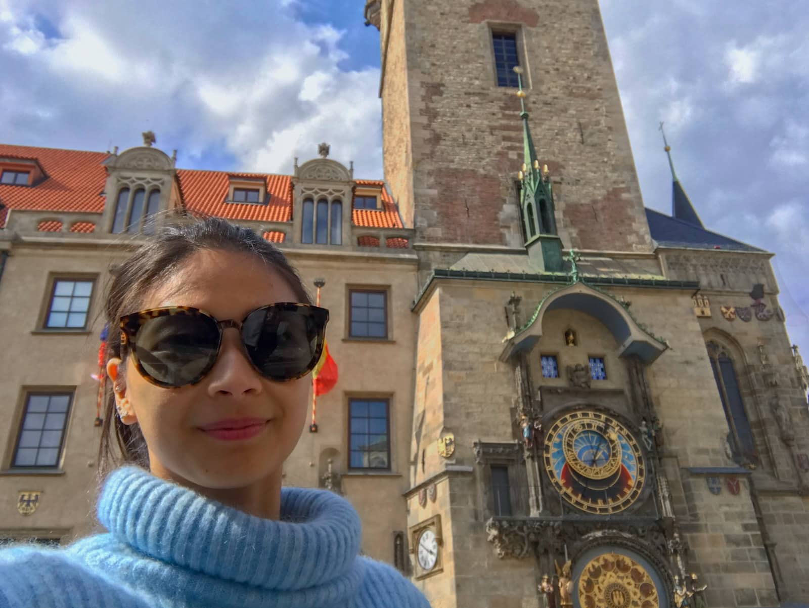 A woman taking a selfie in front of the Astronomical Clock in Prague. She has big round sunglasses on.