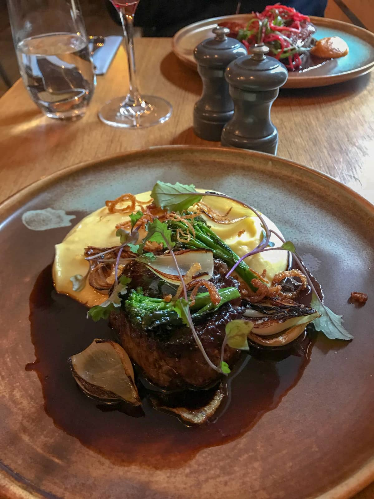 A brown round plate with a cooked steak on it and mashed potato, garnished with onion and broccoli. In the background are salt and pepper shakers and someone else’s meal