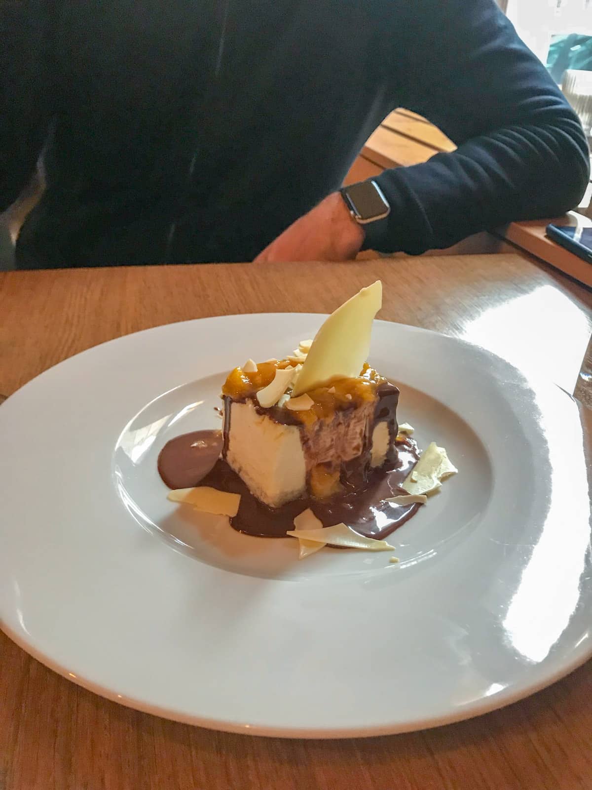 A white plate on a table with a cake on it, dressed with chocolate sauce and white chocolate shards serving as decoration