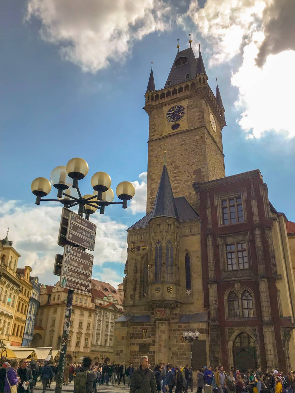 A view of a clock tower in a town square, seen from below. In the foreground is a large signpost with various signs on it and globe lamps on top of the sign