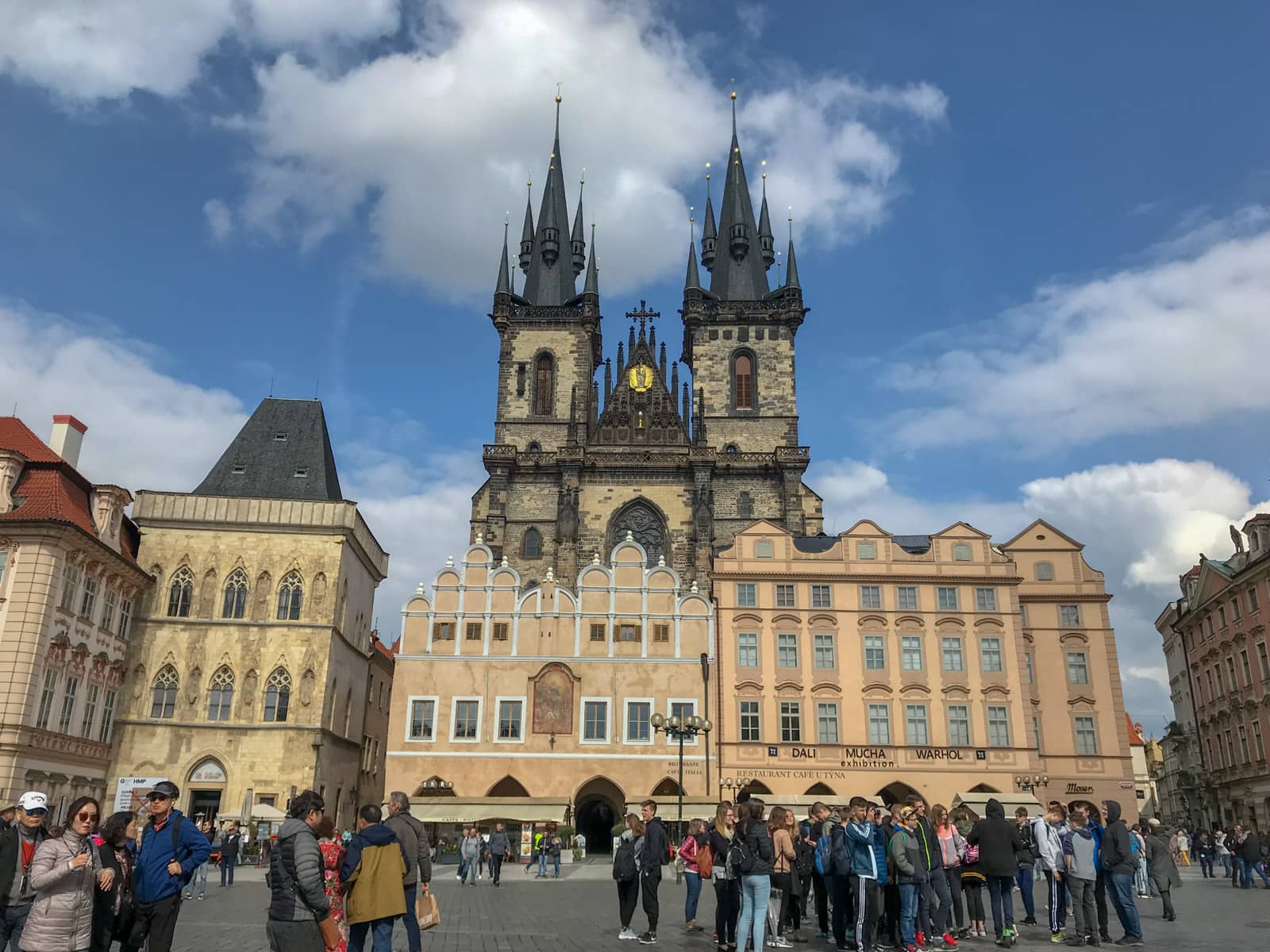 A town square that looks busy with people, with an old building in the background, and a taller, darker building with two towers behind it