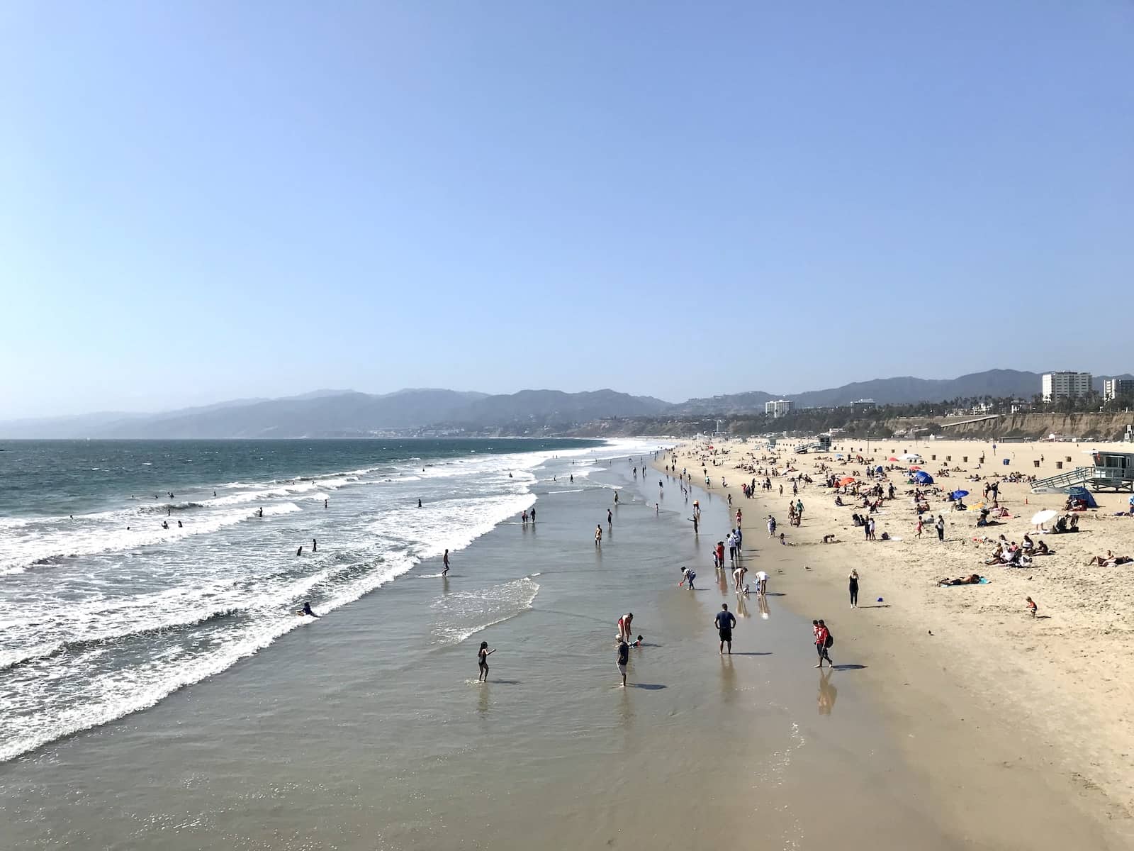 A yellow sand beach by the ocean, with many people on the sand, sitting, standing and enjoying the water and the sand.