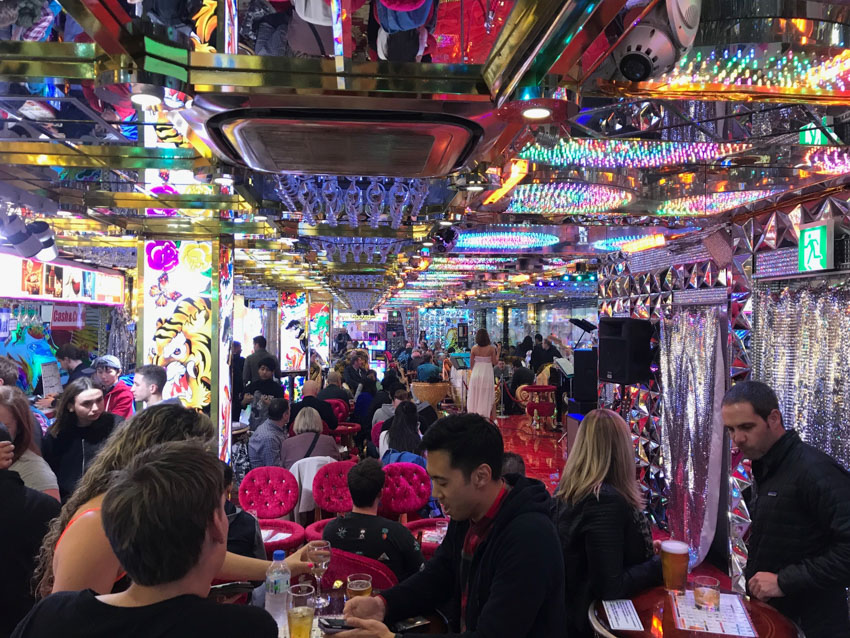 A cramped room with many people standing at small round tables with alcoholic drinks. Some pink velvet chairs surround tables at sitting height. Square pillars inside the room display colourful art. The ceiling has gold accents and is reflective, and also has arrays of colourful LED lights.