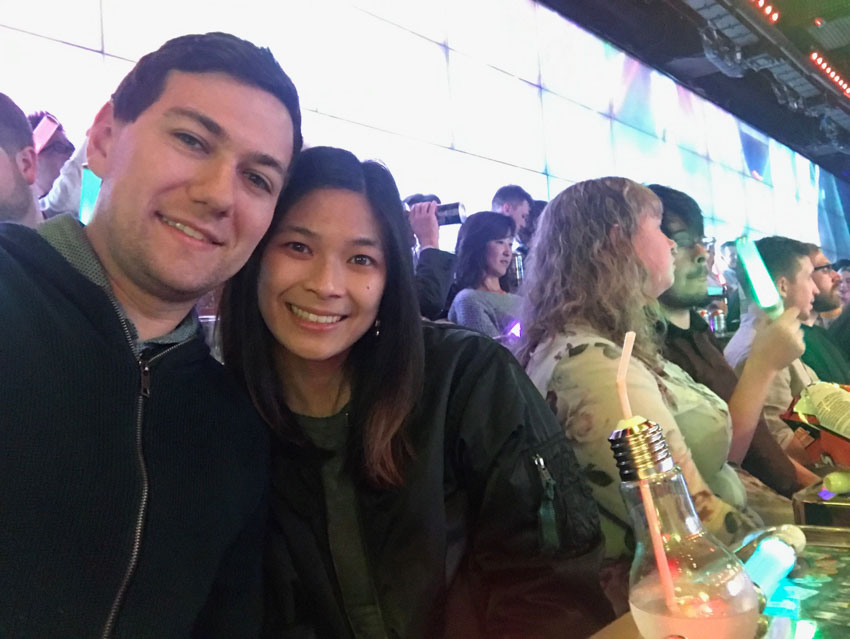 A boy and girl smiling. There are some people seated in the background. On the table in front of the boy and girl is a light bulb shaped bottle with some liquid in it and a straw through the top.